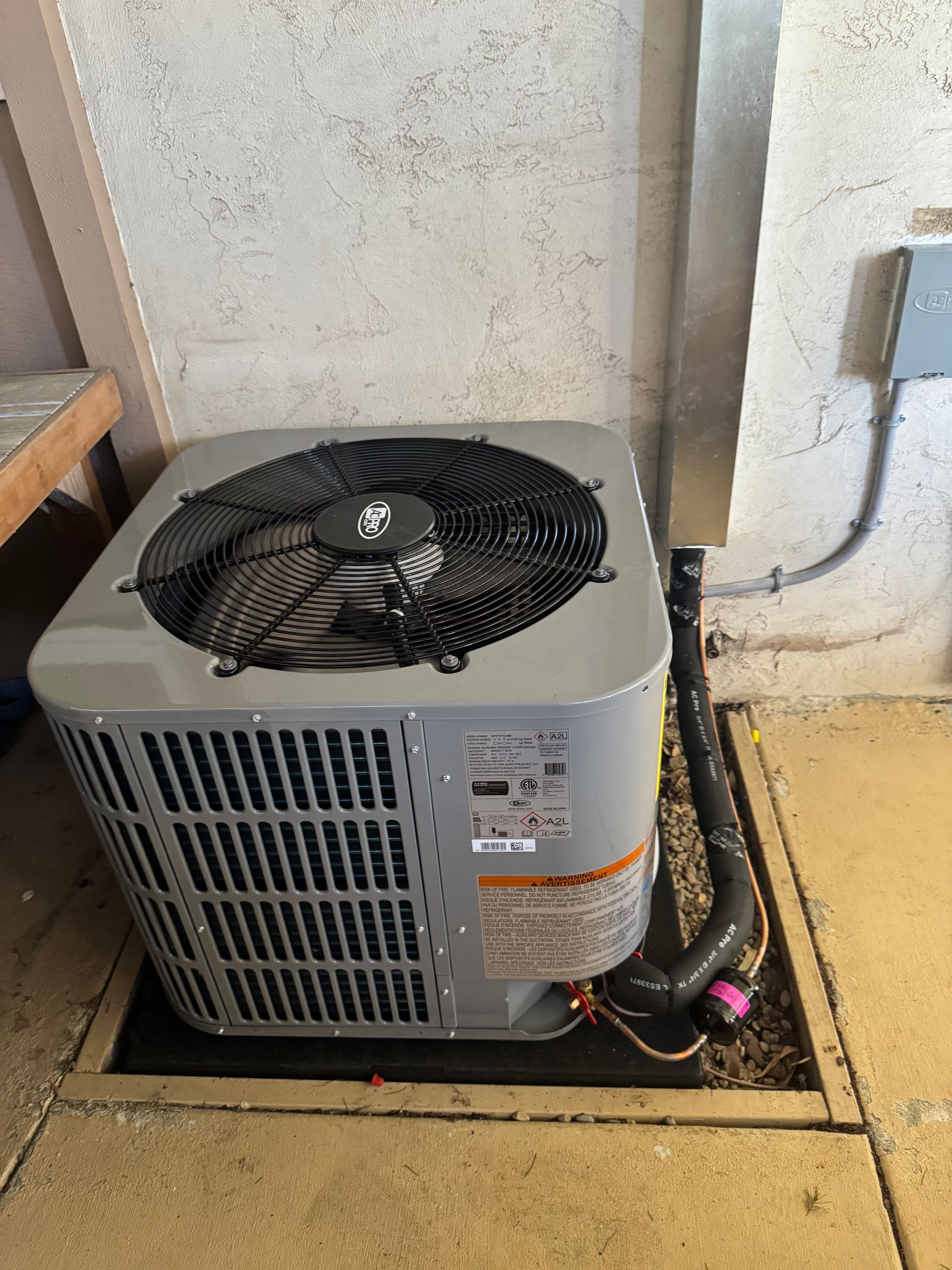 Outdoor air conditioner unit beside a wall, with a fan top and visible pipes on concrete ground.