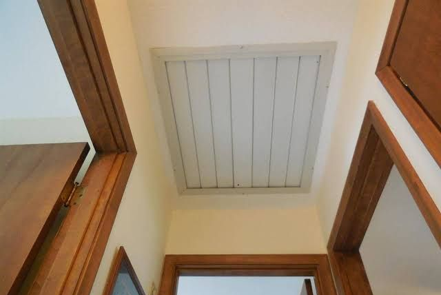 White attic hatch in a beige hallway ceiling framed by wooden door trim