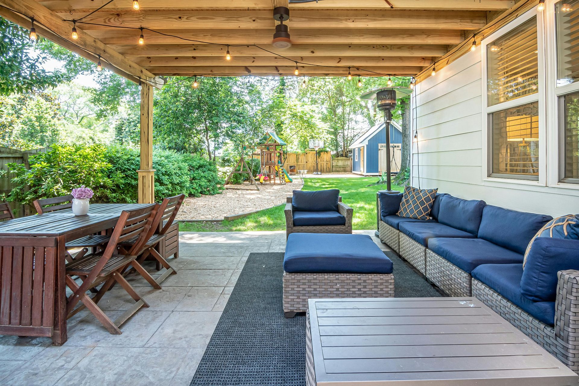 A covered backyard patio with a wooden dining set, a blue sectional sofa, and a matching ottoman on a dark rug.
