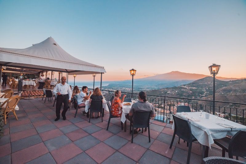 Ristorante con terrazza al tramonto, con vista sulla città, tavoli per cenare e panorama montano in lontananza.