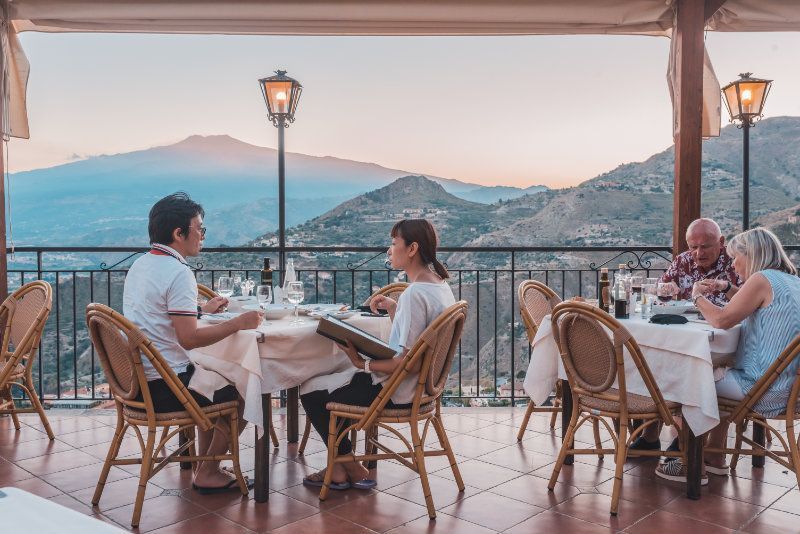 Persone che cenano su una terrazza con vista sulle montagne e sul porto al tramonto.