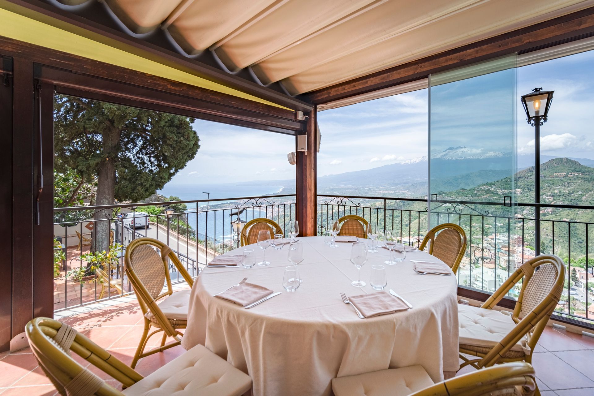 Tavolo da pranzo in terrazza con tovaglia bianca, sedie in vimini e vista sulle montagne e sul mare oltre le ringhiere in vetro.