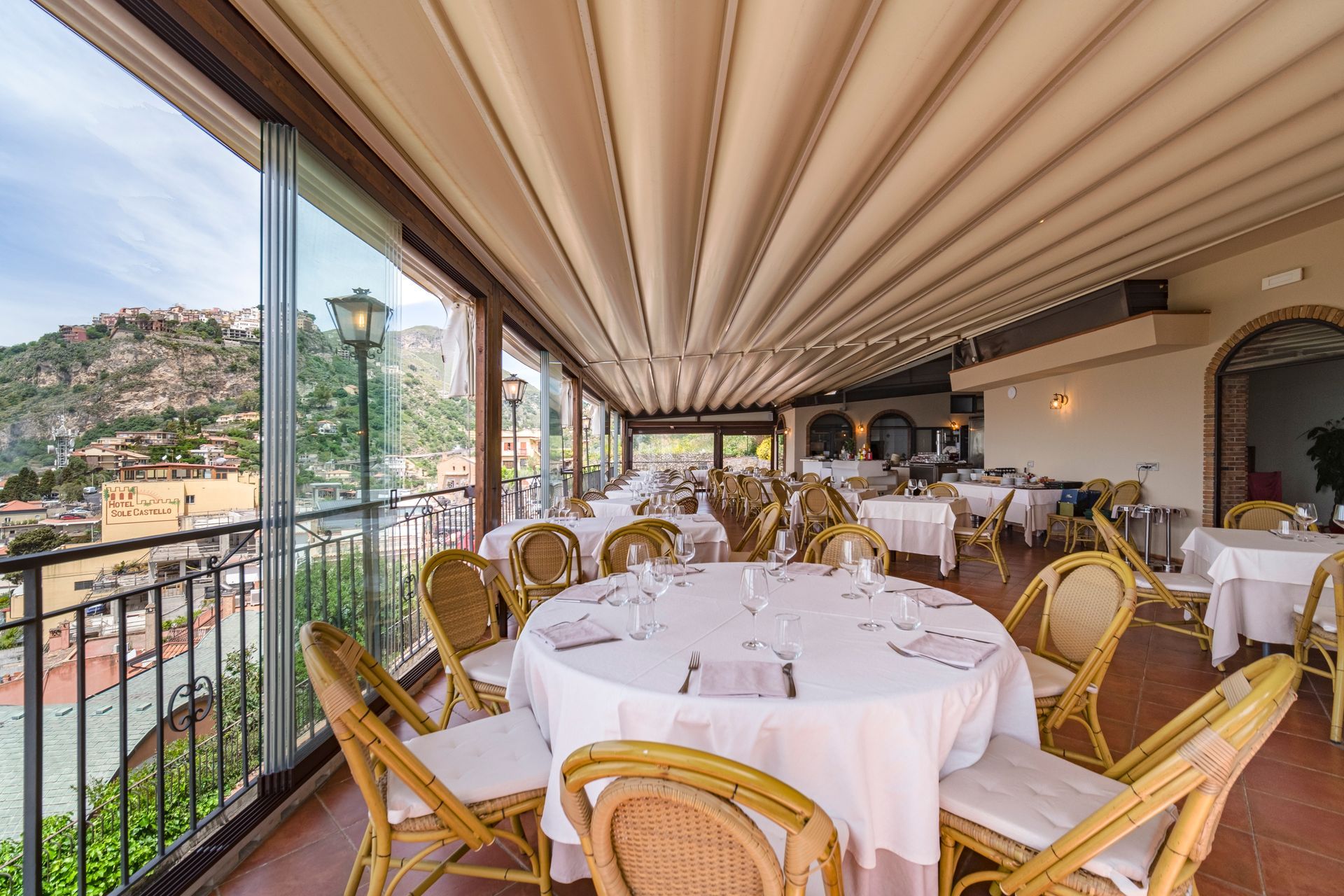 Elegante zona pranzo in terrazza con tovaglie bianche, sedie in vimini e vista sulle montagne attraverso pareti di vetro.