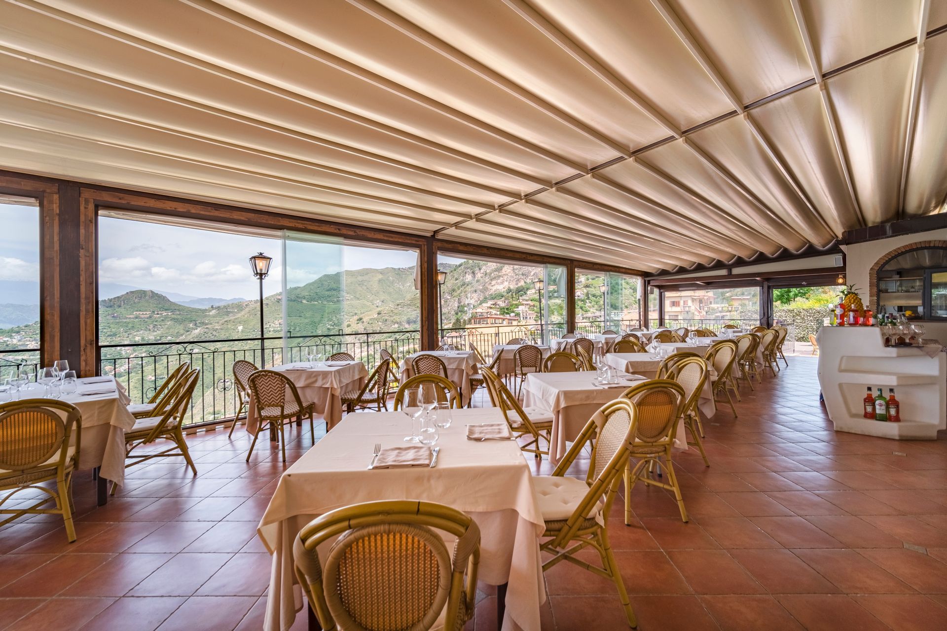 Ristorante all'aperto con sedie in vimini, tovaglie bianche e vista sulle montagne al tramonto.