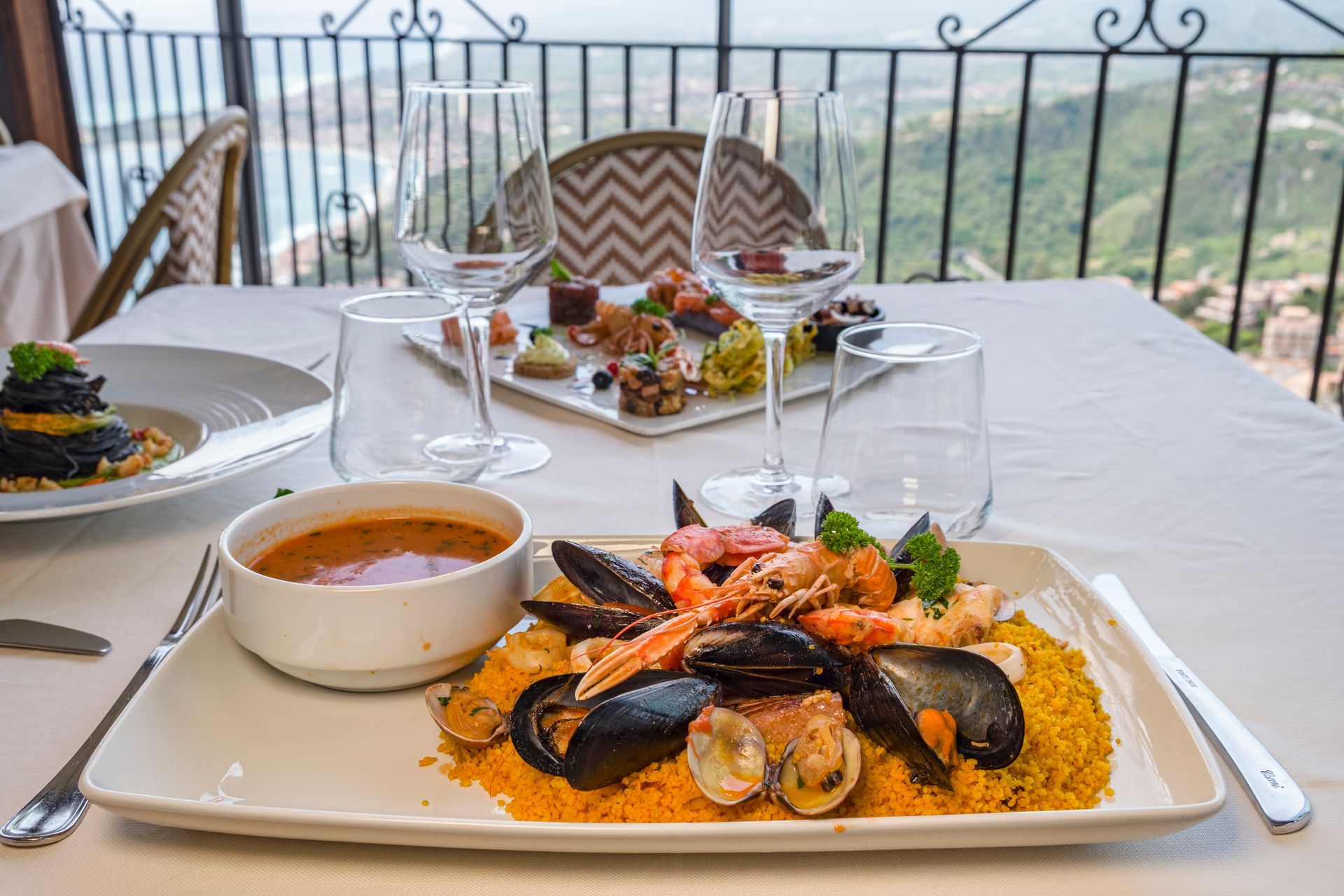 Tavolo all'aperto con piatto di frutti di mare, zuppa e bevande su un balcone con vista sull'acqua.