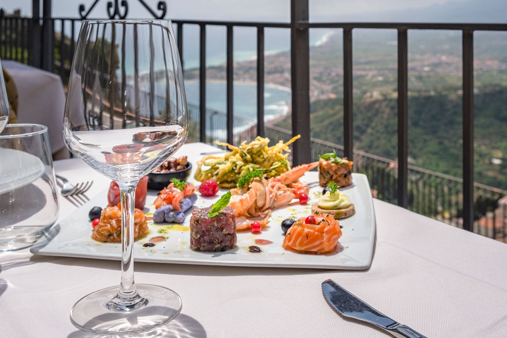 Elegante tavolo da pranzo all'aperto con antipasti gourmet e vino, con vista su una suggestiva costa.