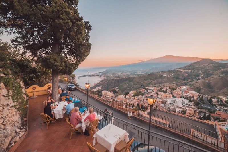 Cenare all'aperto con vista su una cittadina costiera e sul mare al tramonto, con le montagne sullo sfondo.