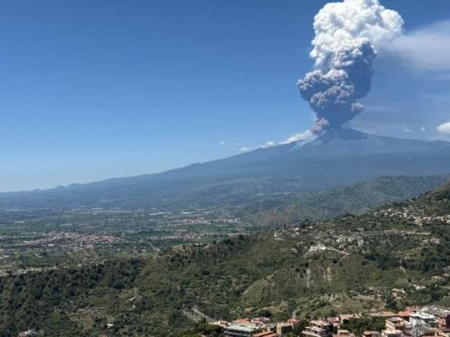 Eruzione vulcanica con un'enorme colonna di cenere che si innalza su una verde vallata e una città lontana, sotto un cielo azzurro e limpido.