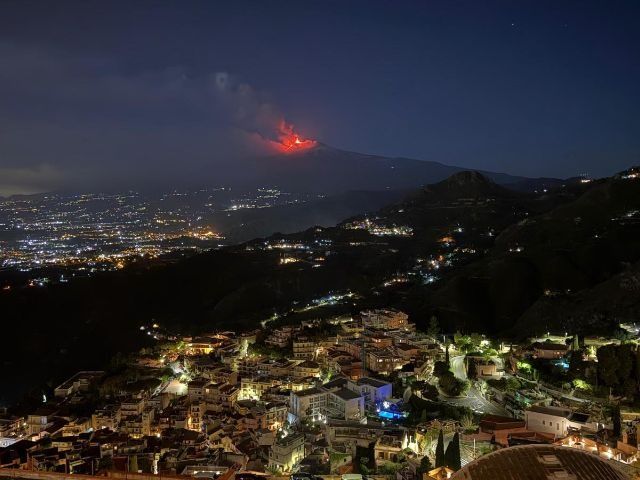 Paesaggio urbano notturno con luci scintillanti e una lontana eruzione vulcanica rossa all'orizzonte.