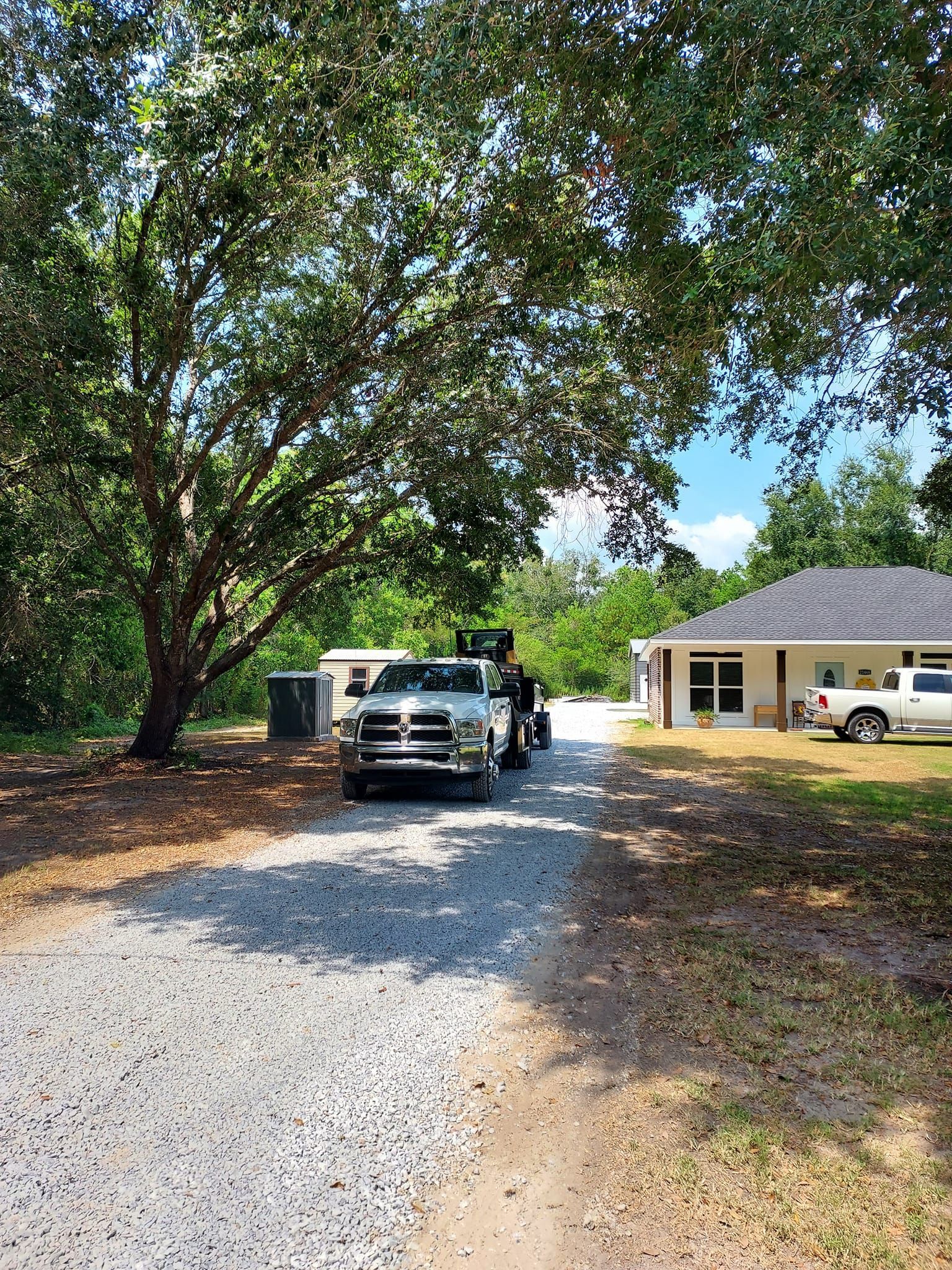 A gravel driveway with a truck and building, shaded by a large tree, on a sunny day.