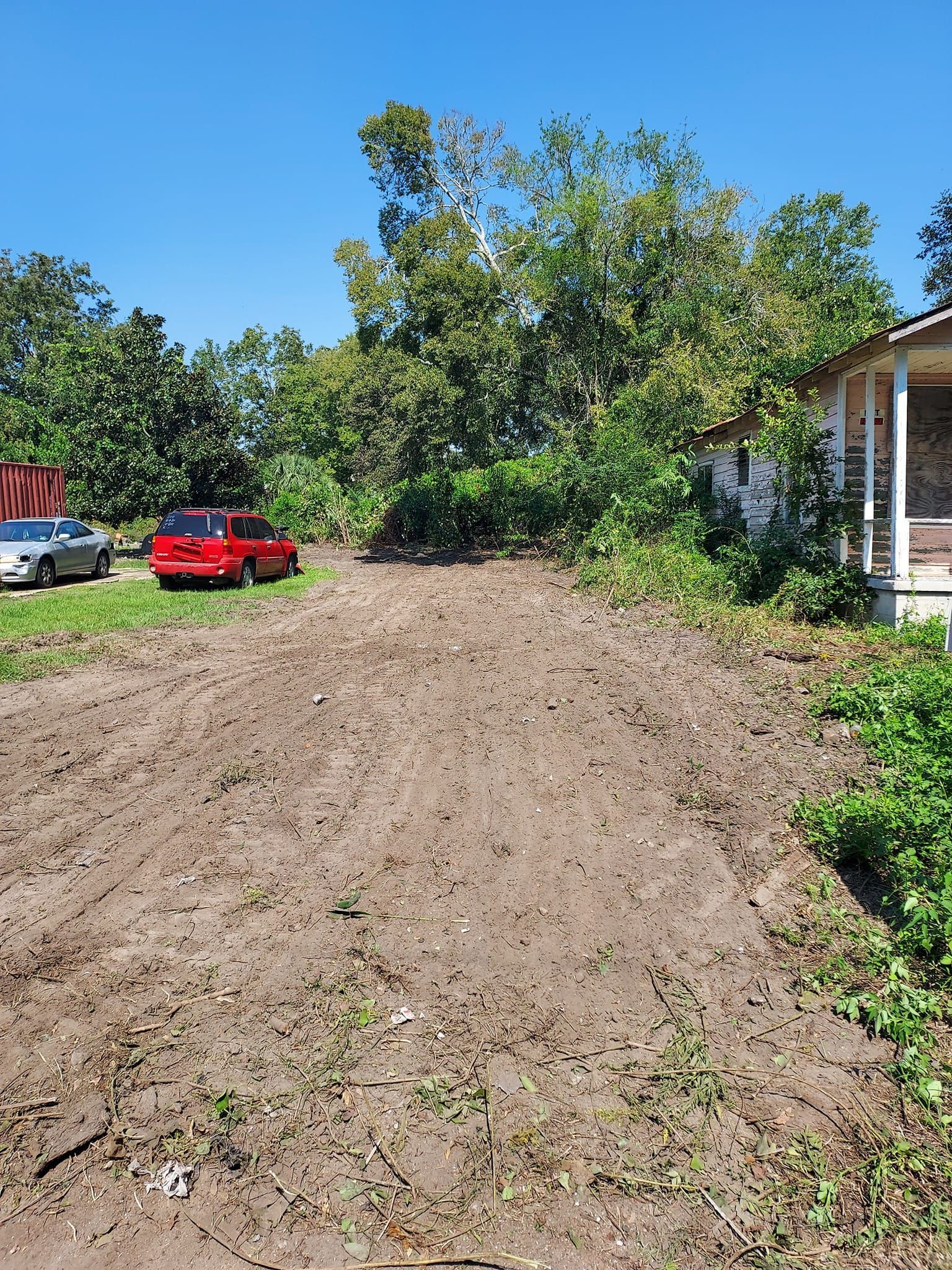 Muddy driveway with cars, leading to a house partially obscured by trees, under a blue sky.