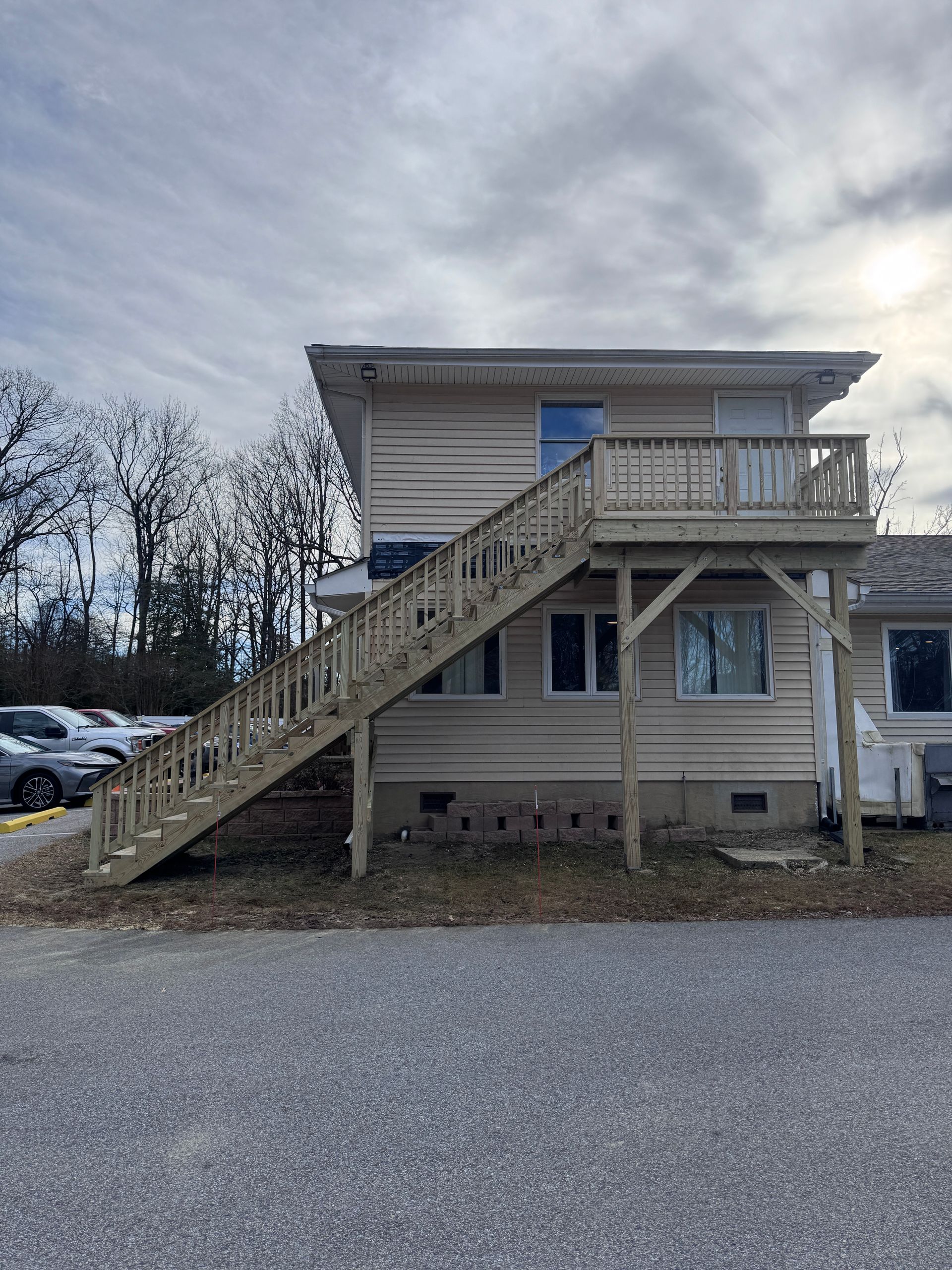 Two-story beige house with an exterior staircase and gravel parking area under a cloudy sky
