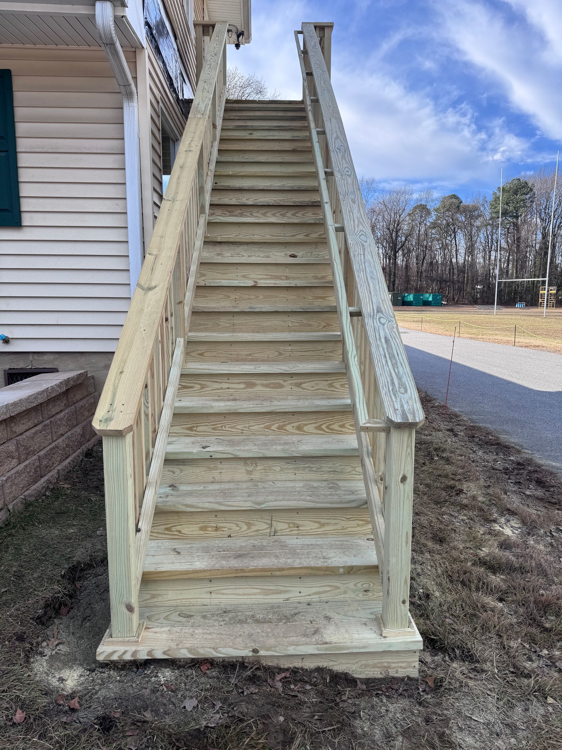 Wooden outdoor staircase with railings beside a house on a grassy yard, under a blue sky