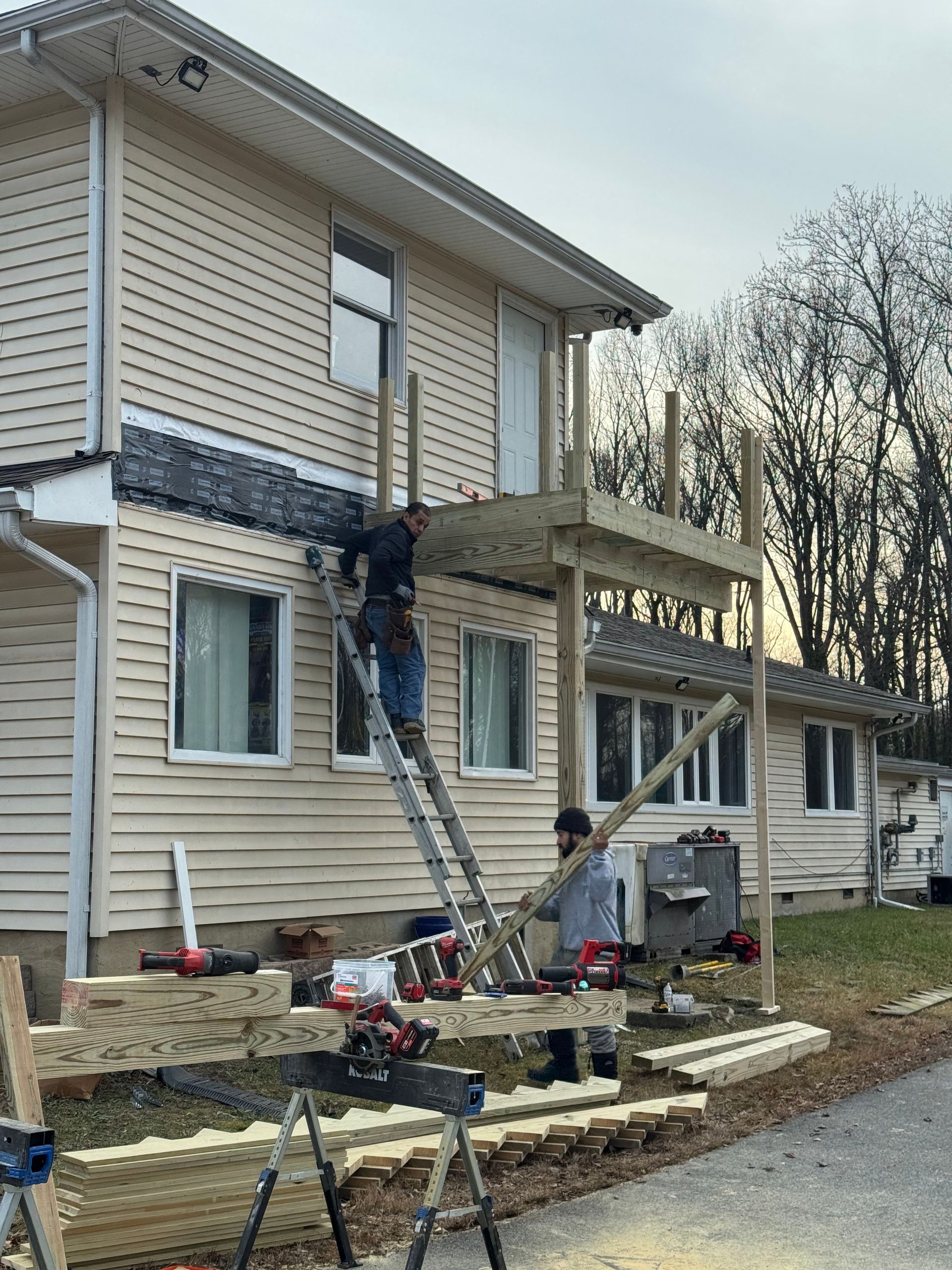 Worker on a ladder repairing siding and a porch on a beige house under construction.