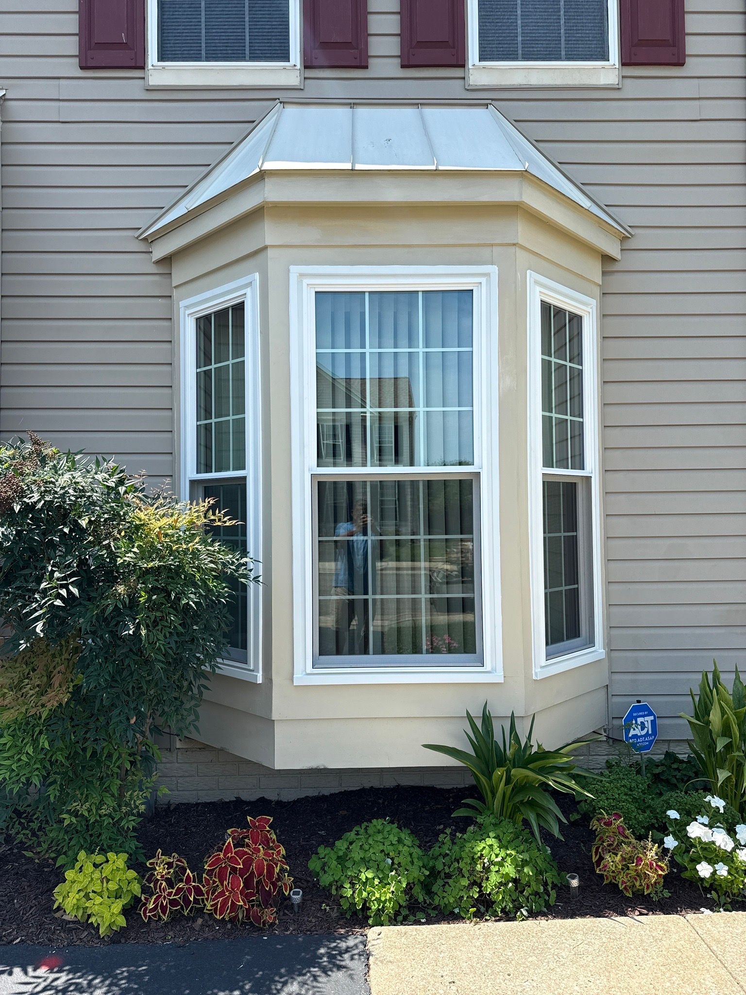 Bay window with white trim on a beige house, framed by shrubs and flowers in a small garden.
