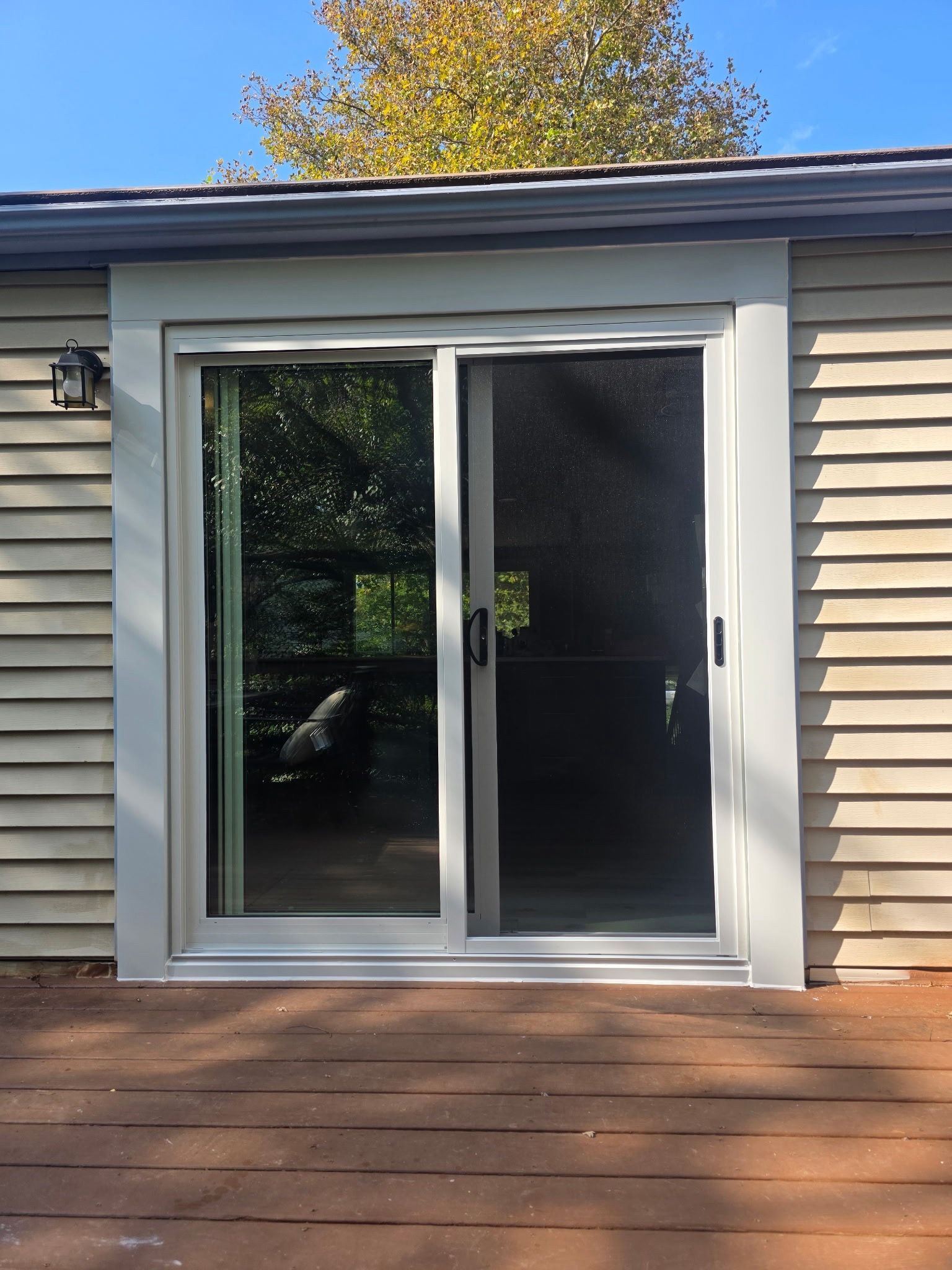 Sliding glass door on a beige house exterior with wooden deck and autumn trees reflected in the glass