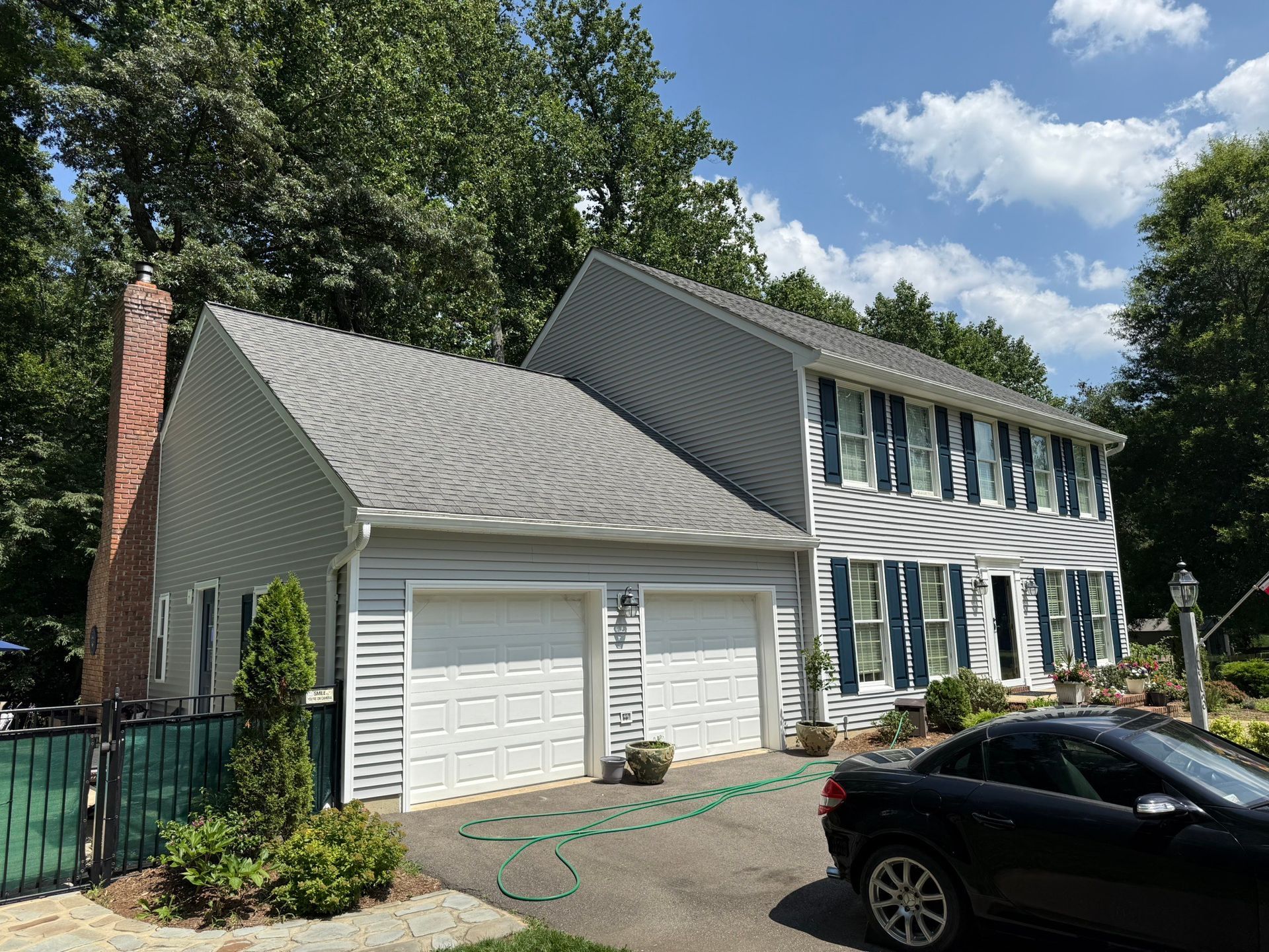 Suburban two-story house with white garage doors, blue-gray siding, and a black car in front under a sunny sky