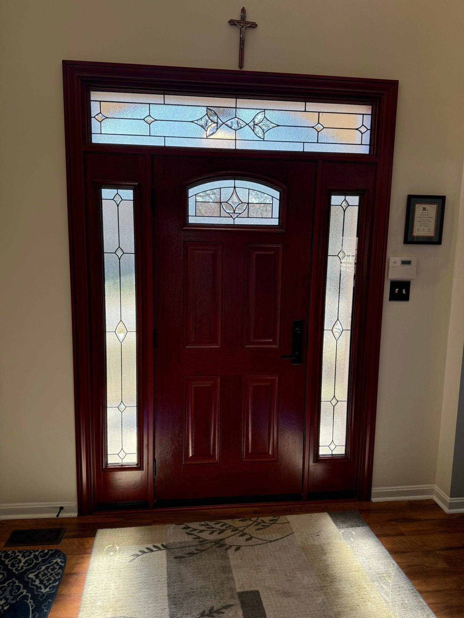 Front door with stained-glass sidelights and transom, beneath a small cross, lit by sunlight.