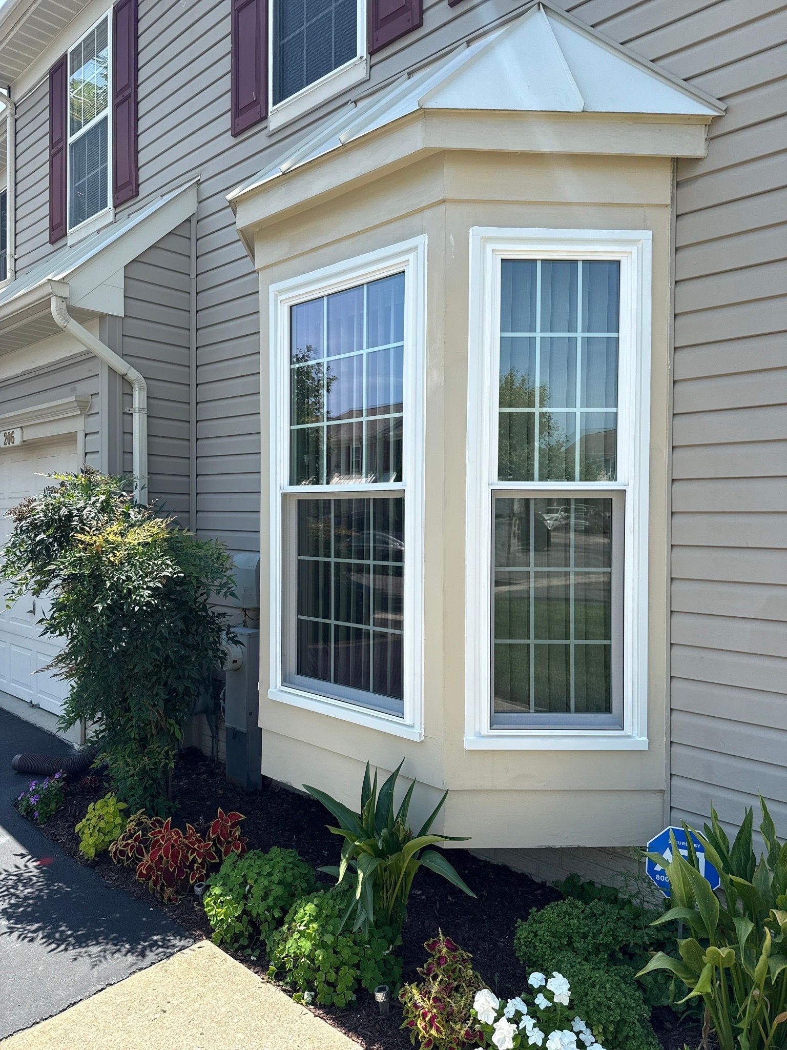 Exterior bay window on a beige house with white trim and small landscaping plants.
