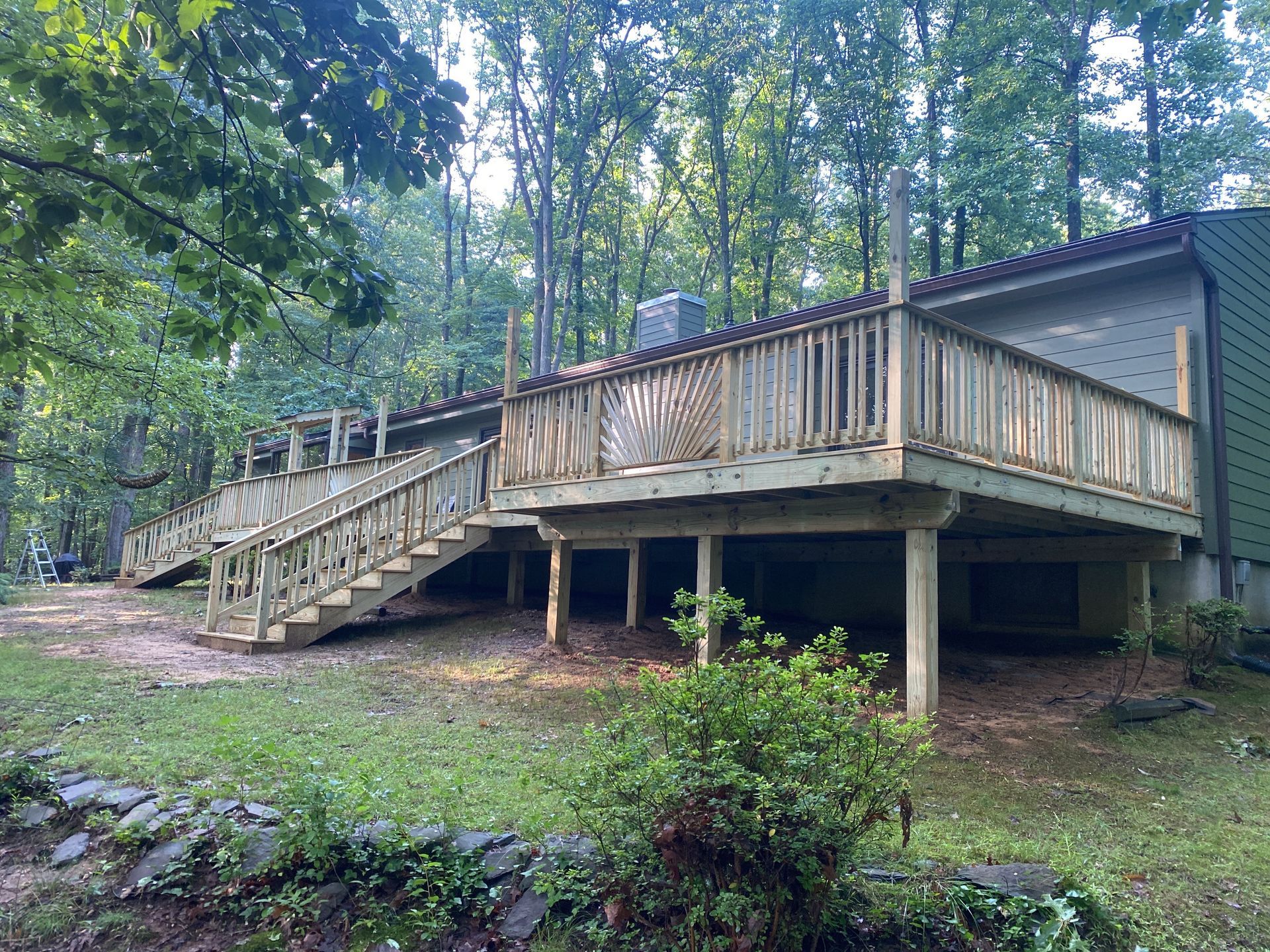 Wooden deck with stairs on stilts in a wooded area, surrounded by trees and greenery