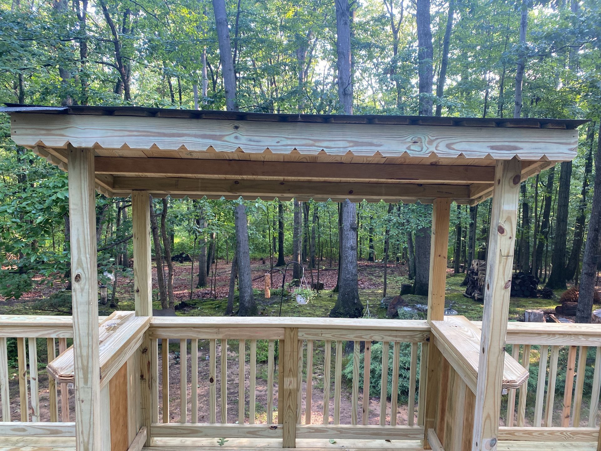 Wooden deck gazebo under construction in a wooded backyard, with railings and unfinished roof beams