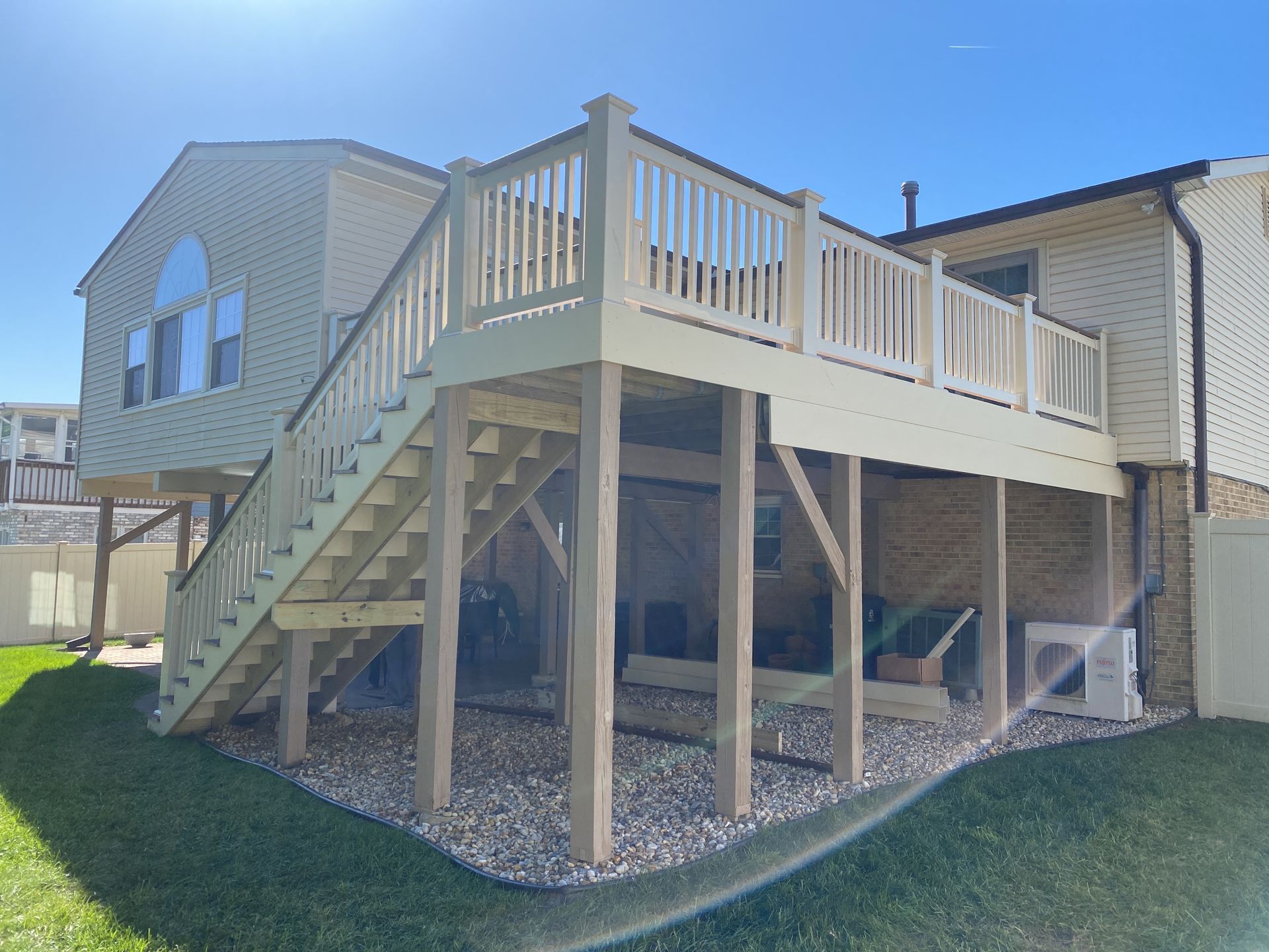 Elevated backyard deck with stairs, white railing, and support posts beside a beige house under a blue sky