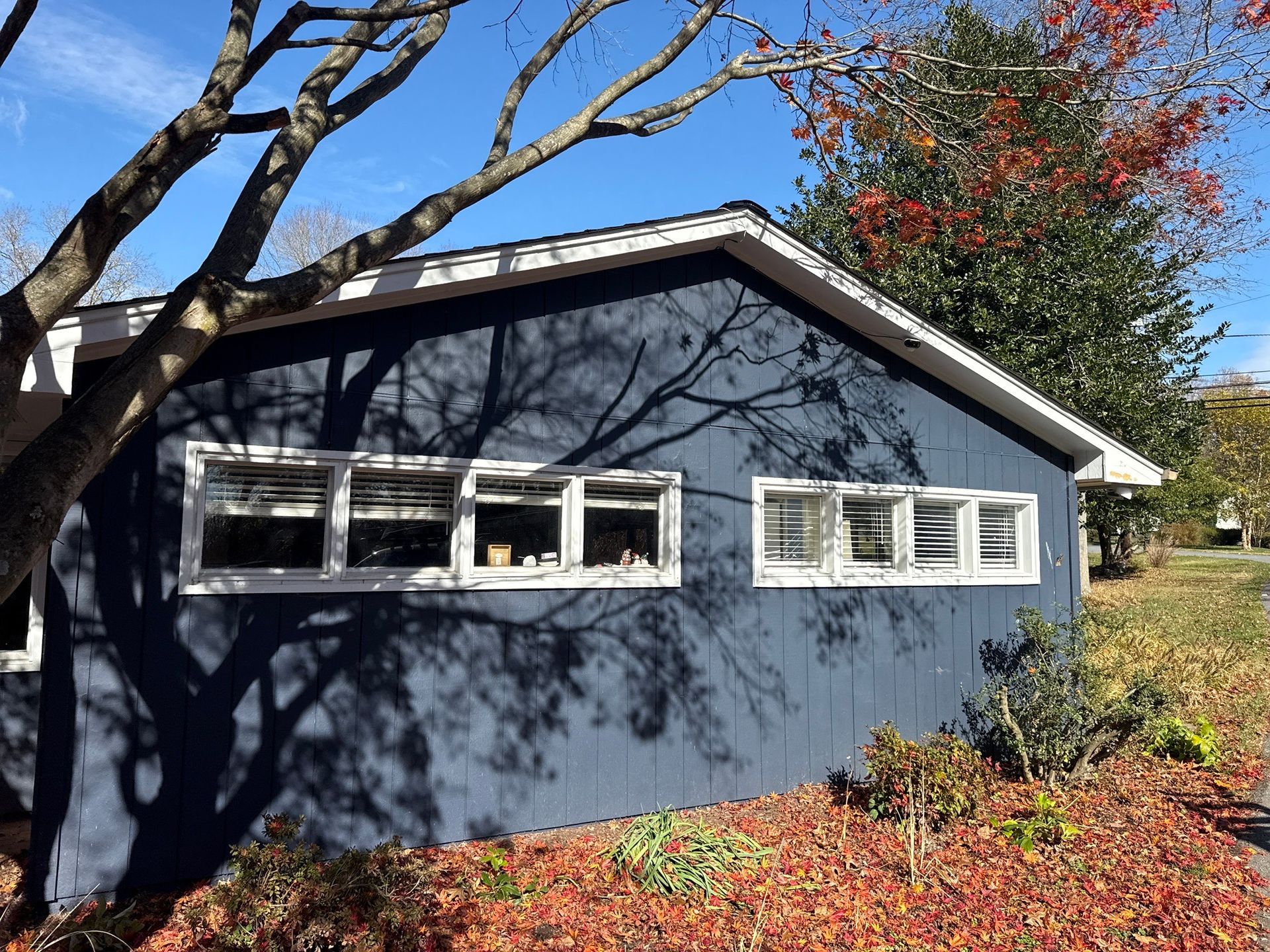 Blue house with white-trimmed windows and a dark roof, partly shaded by trees in autumn.