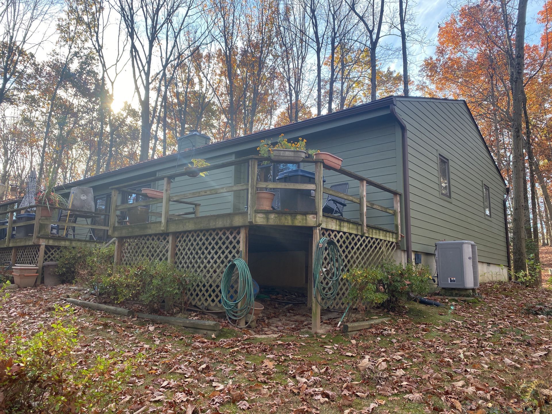 A gray house with a wooden deck and lattice skirting, surrounded by autumn trees and fallen leaves.