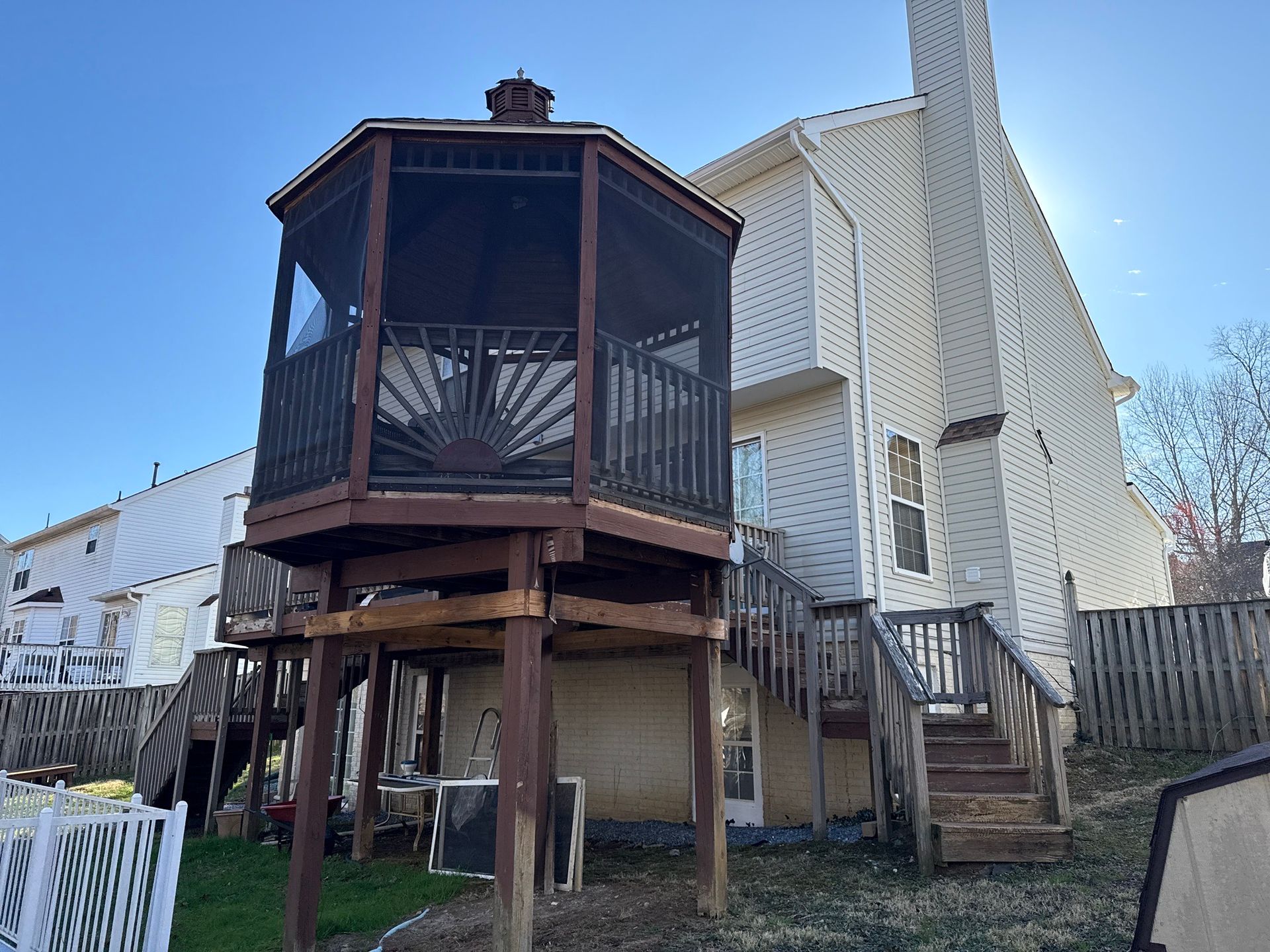 A raised gazebo-like deck with lattice sides stands in a backyard beside a white house and wooden fence.