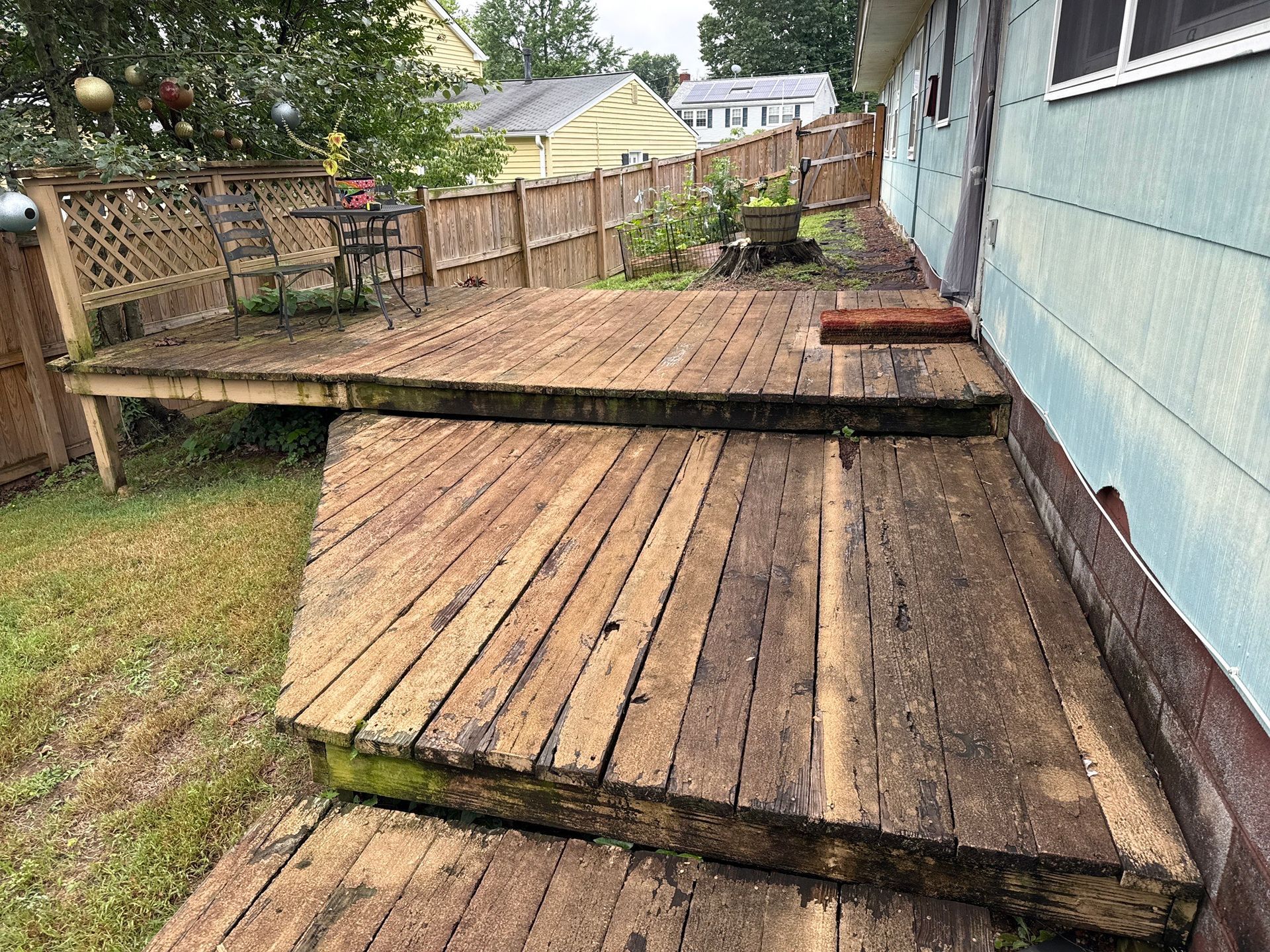 Weathered wooden backyard deck beside a blue house, with a raised walkway and fenced yard.