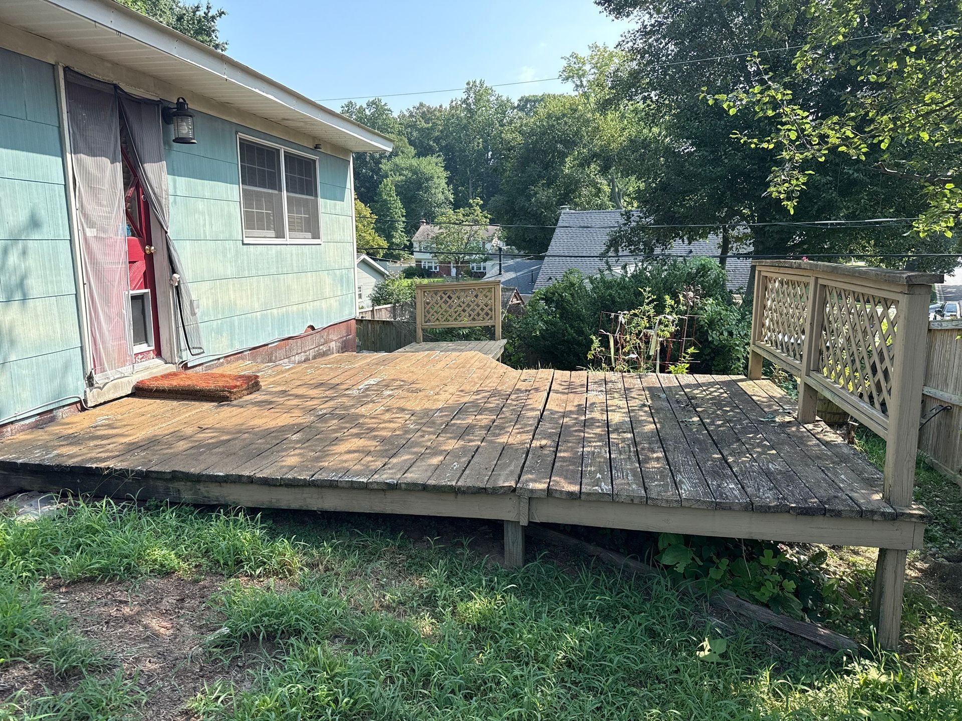 Weathered wooden deck beside a house, with a railing and green trees in the background.