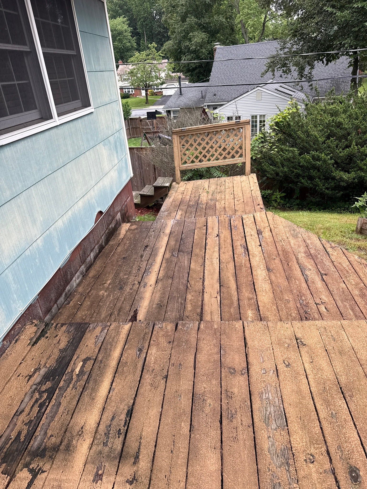 Wooden deck and ramp beside a blue house, leading to a fenced backyard with trees.