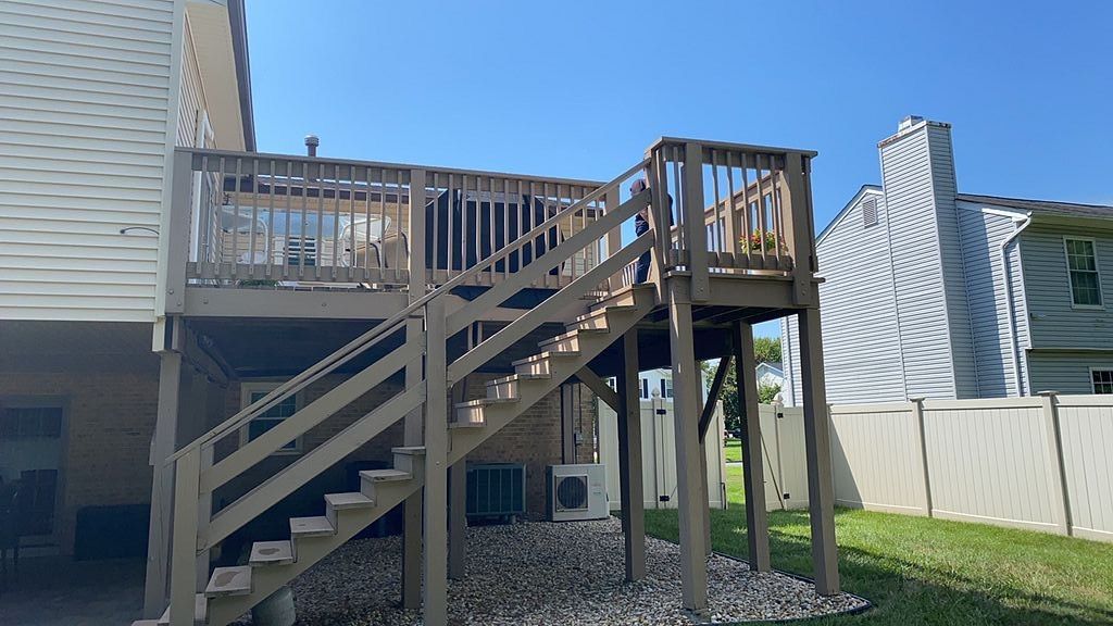 Wooden raised deck with stairs beside a house and fenced backyard under a blue sky