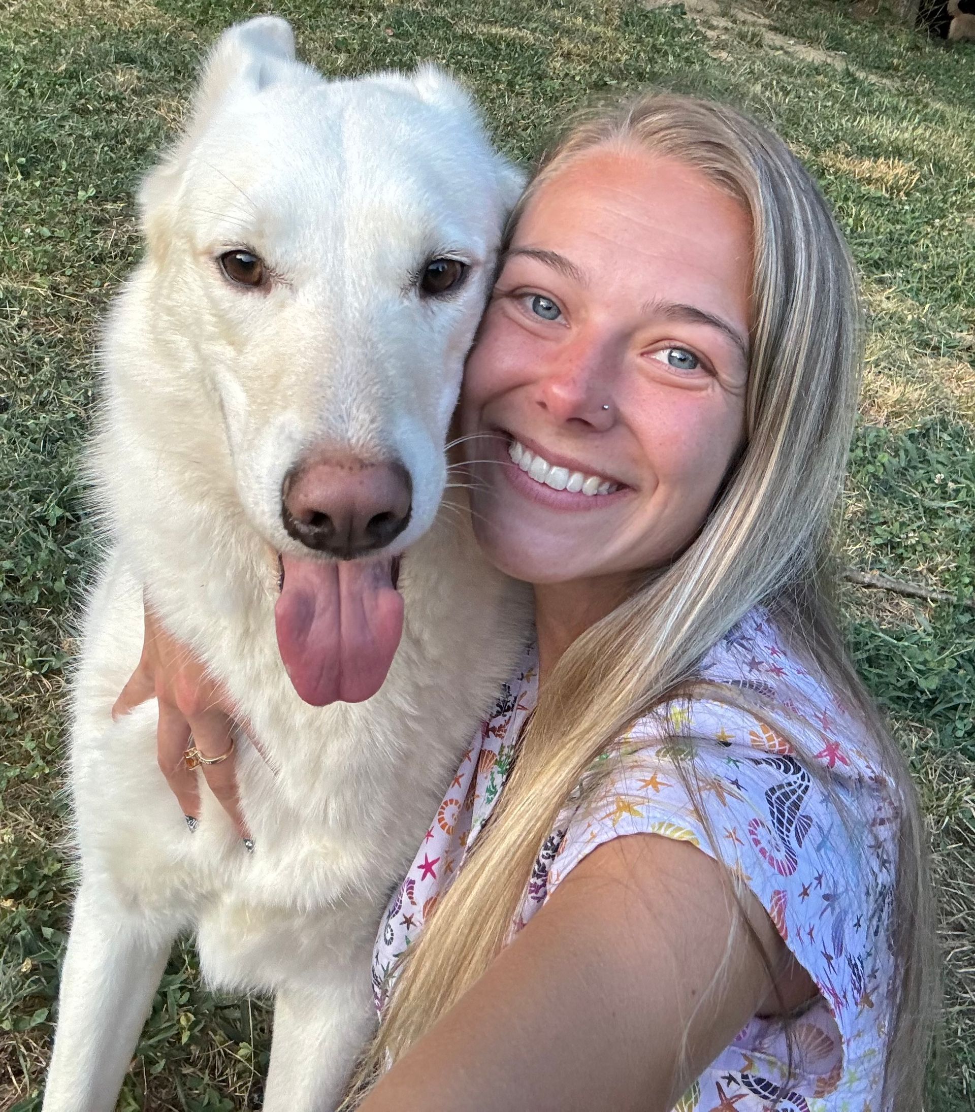 Woman smiling beside a white dog with its tongue out, both close to the camera outdoors.