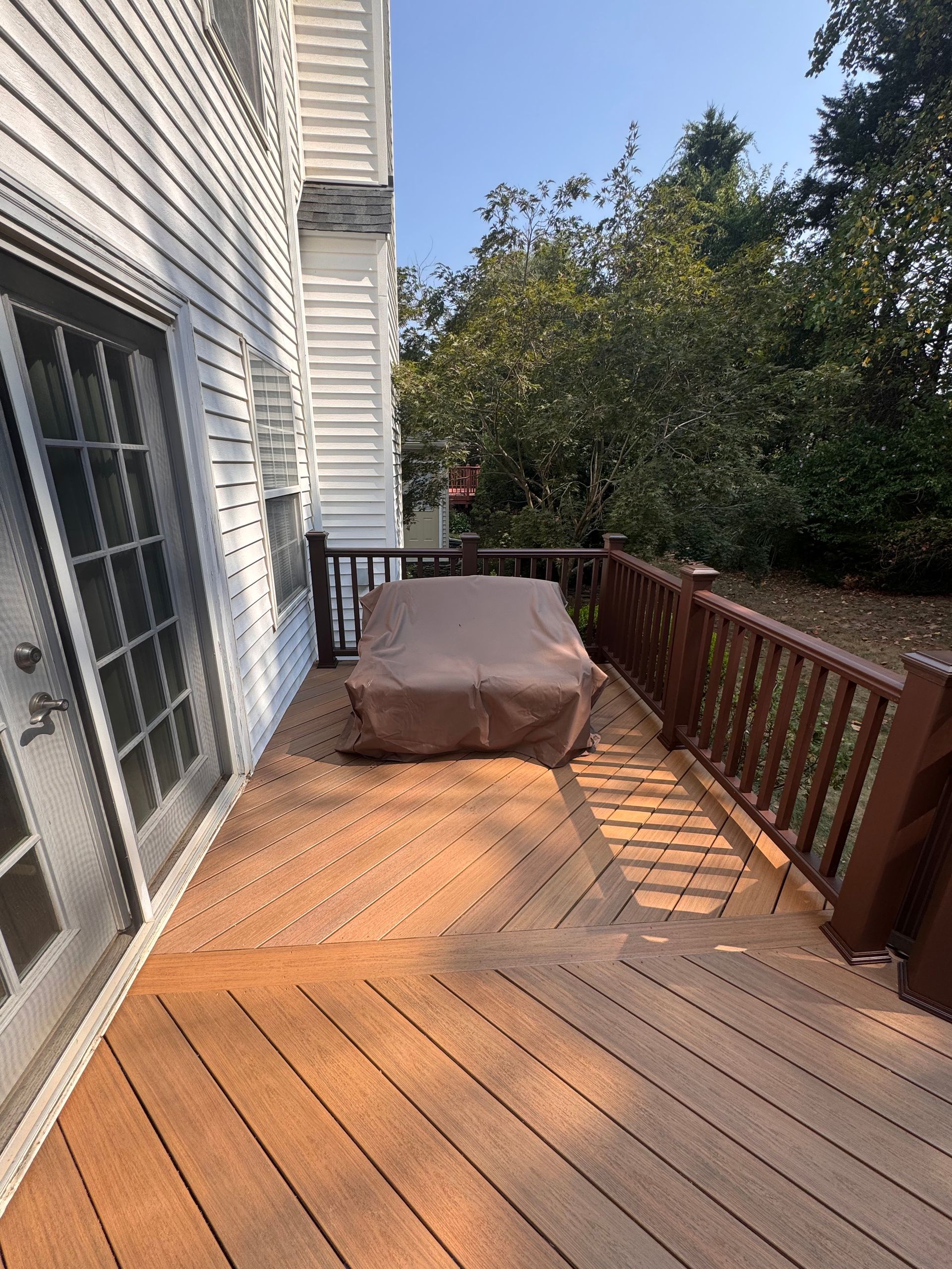 Wooden backyard deck beside a white house, with a covered grill and railing overlooking trees.