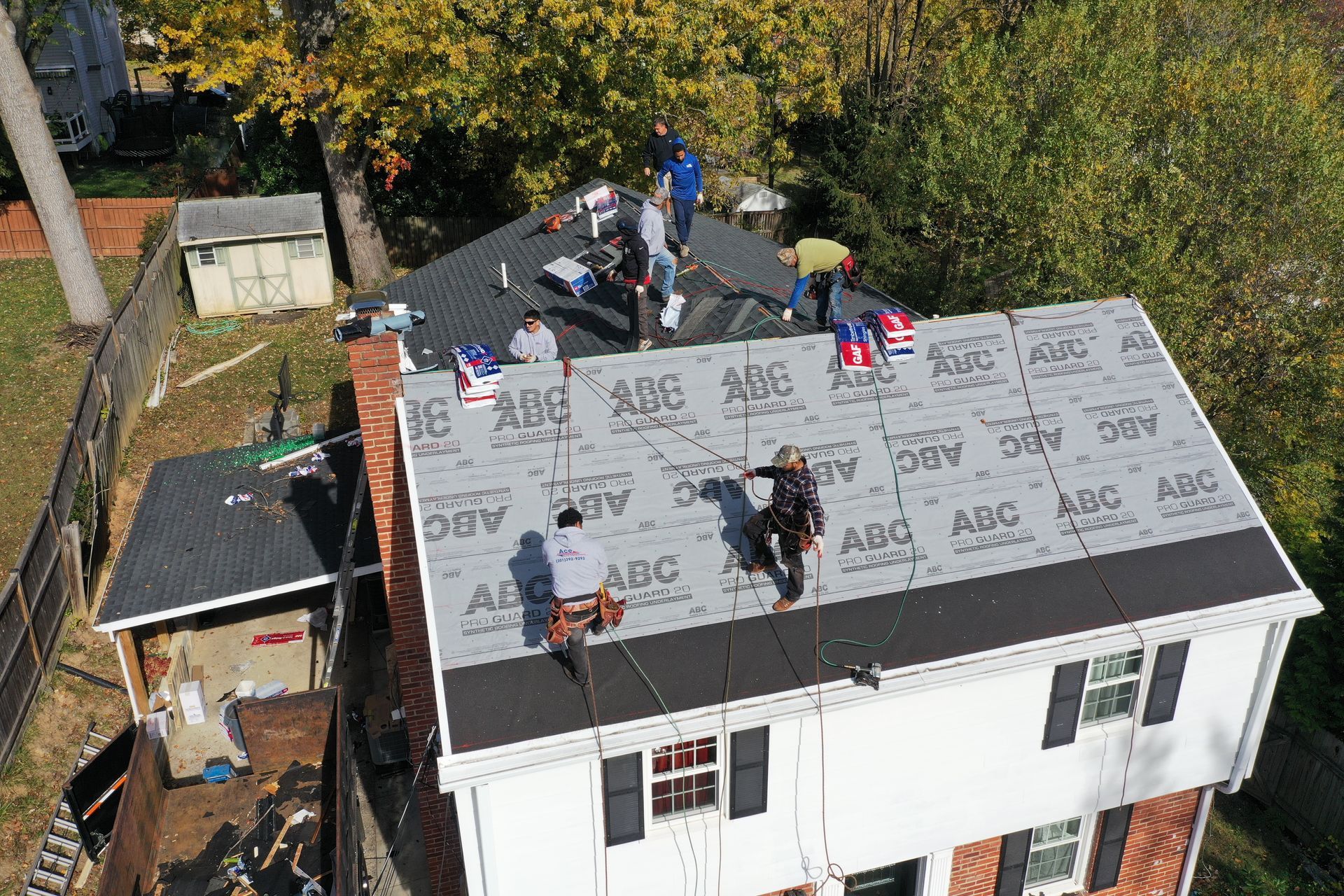 Roofers installing shingles on a white house roof with gray underlayment and trees nearby.