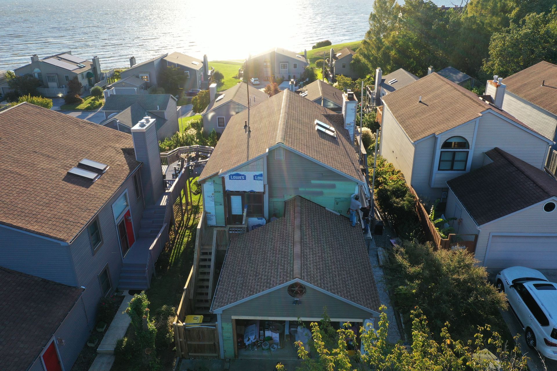 Aerial view of houses by a sunlit waterfront, with roofs, trees, and parked cars.