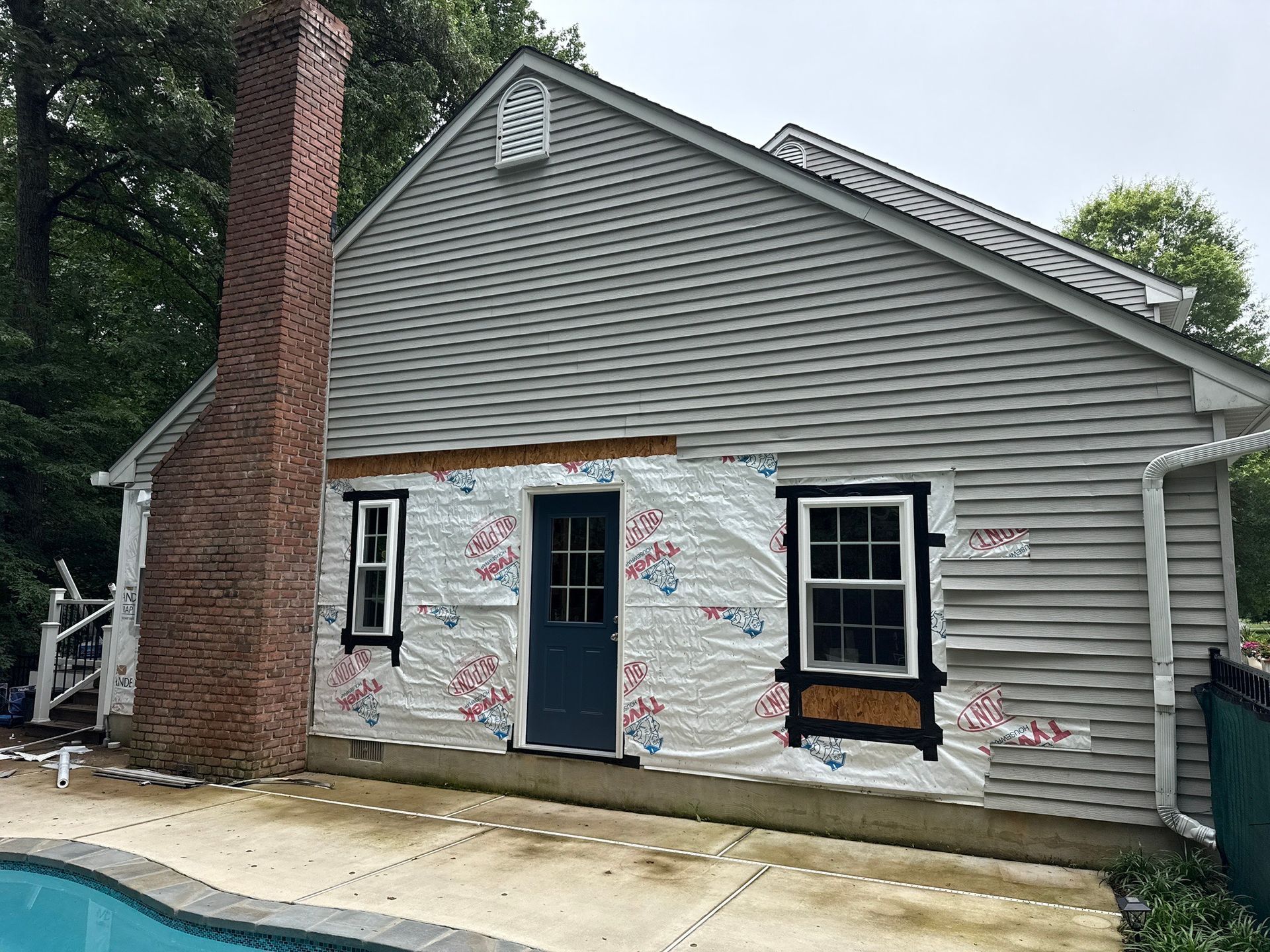 Gray house exterior with brick chimney and boarded windows covered in spray paint, under renovation