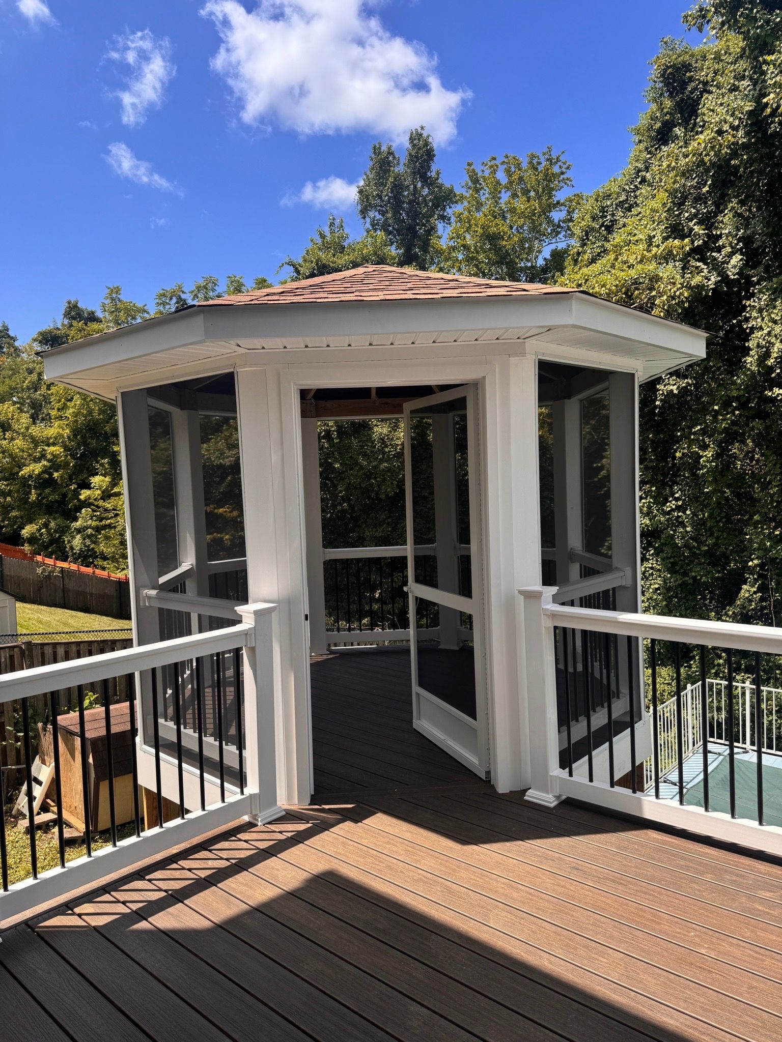 White gazebo with open doors on a wooden deck, surrounded by trees under a blue sky.