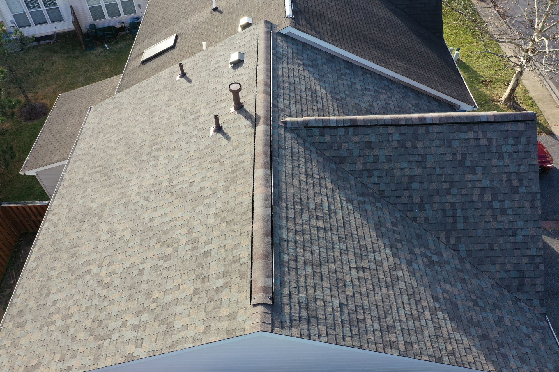 Aerial view of a shingled house roof with vents and a chimney, bordered by a driveway and yard.