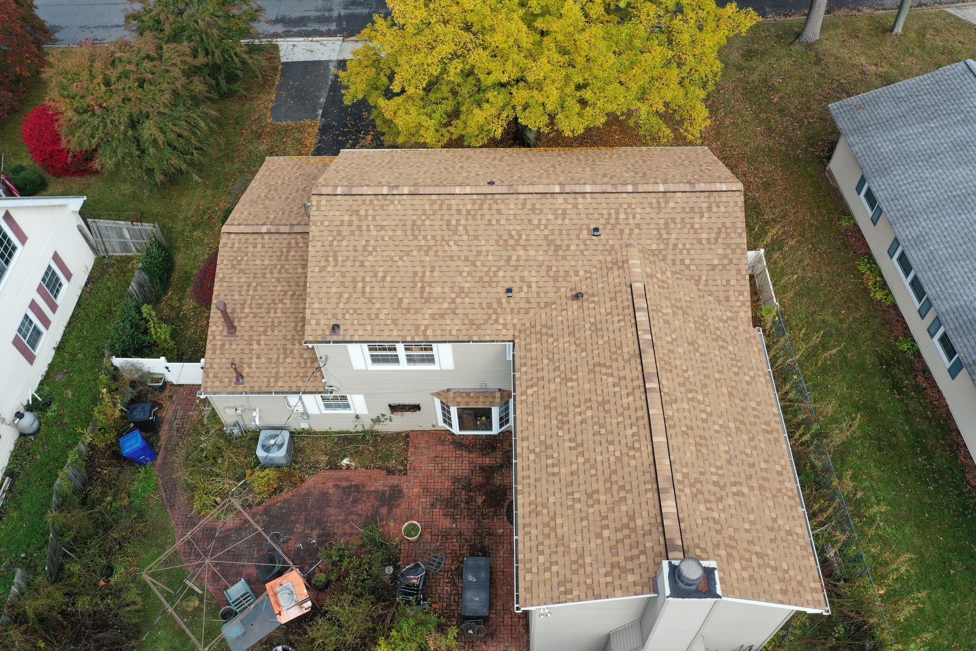 Aerial view of a tan-roofed house with a backyard, surrounded by trees and neighboring homes.