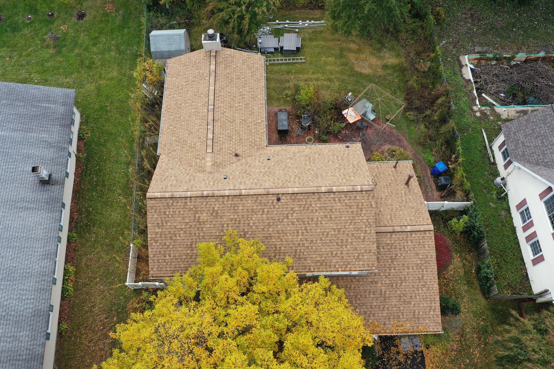 Aerial view of a tan shingled house with a large roof and surrounding trees and neighboring homes.