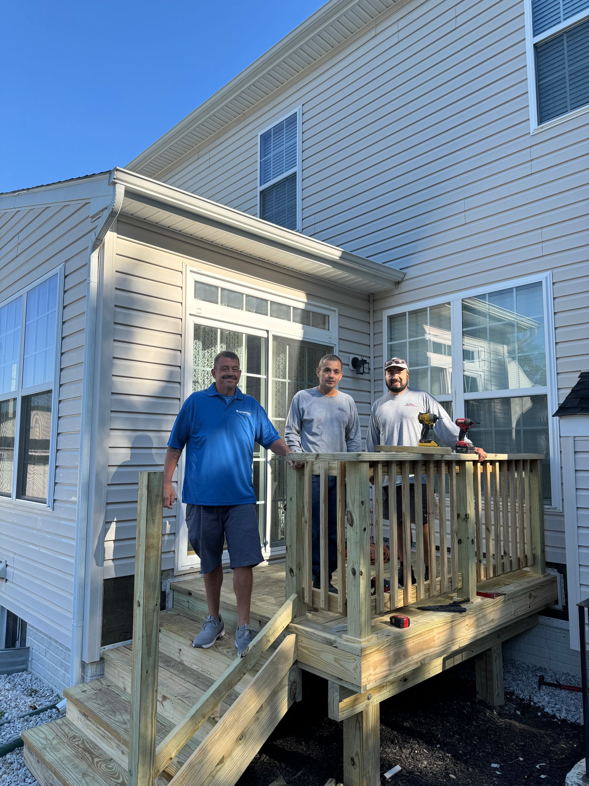 Three people on a small wooden deck beside a light gray house on a sunny day