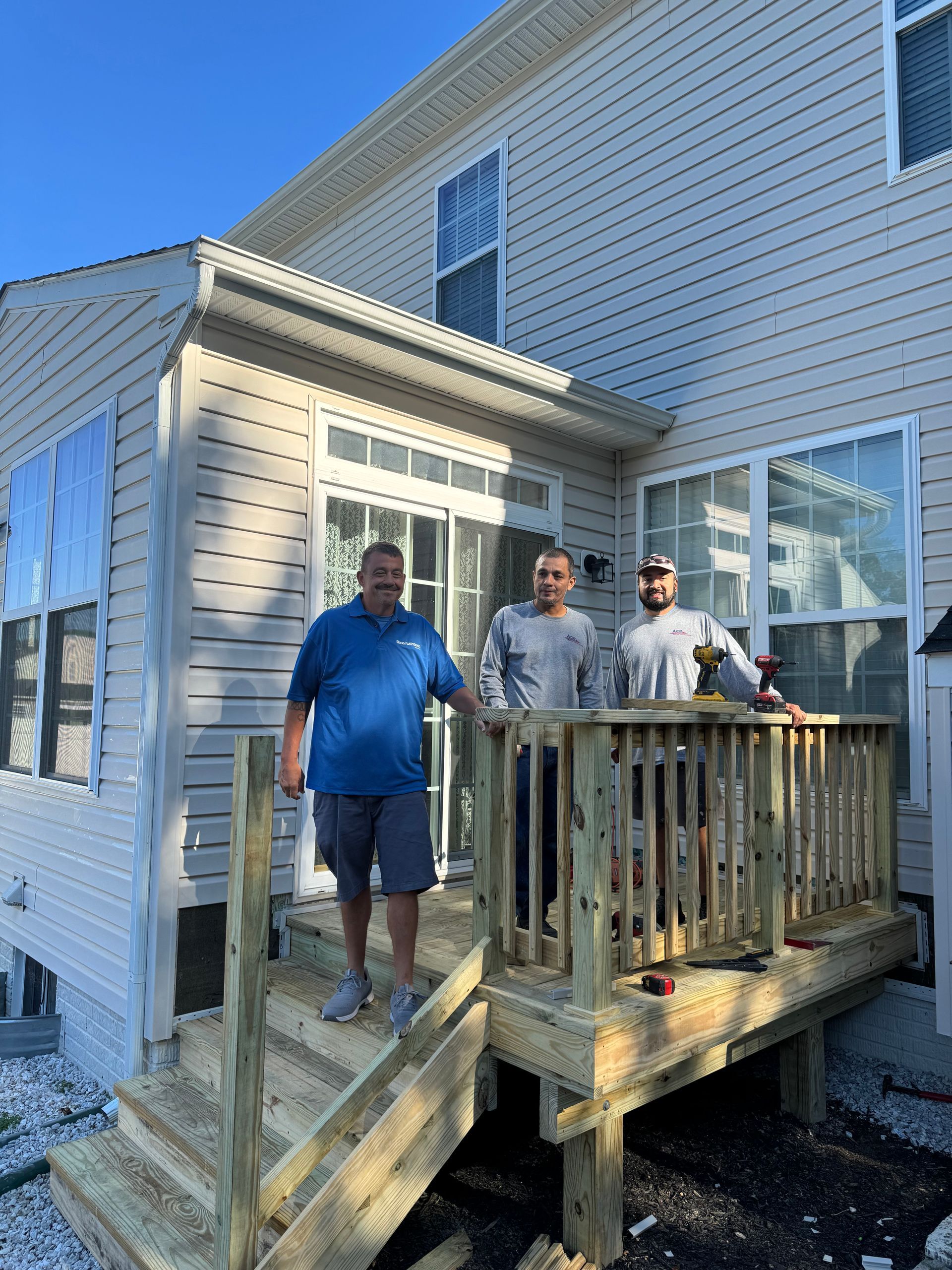 Three people standing on a small wooden deck beside a house in bright daylight