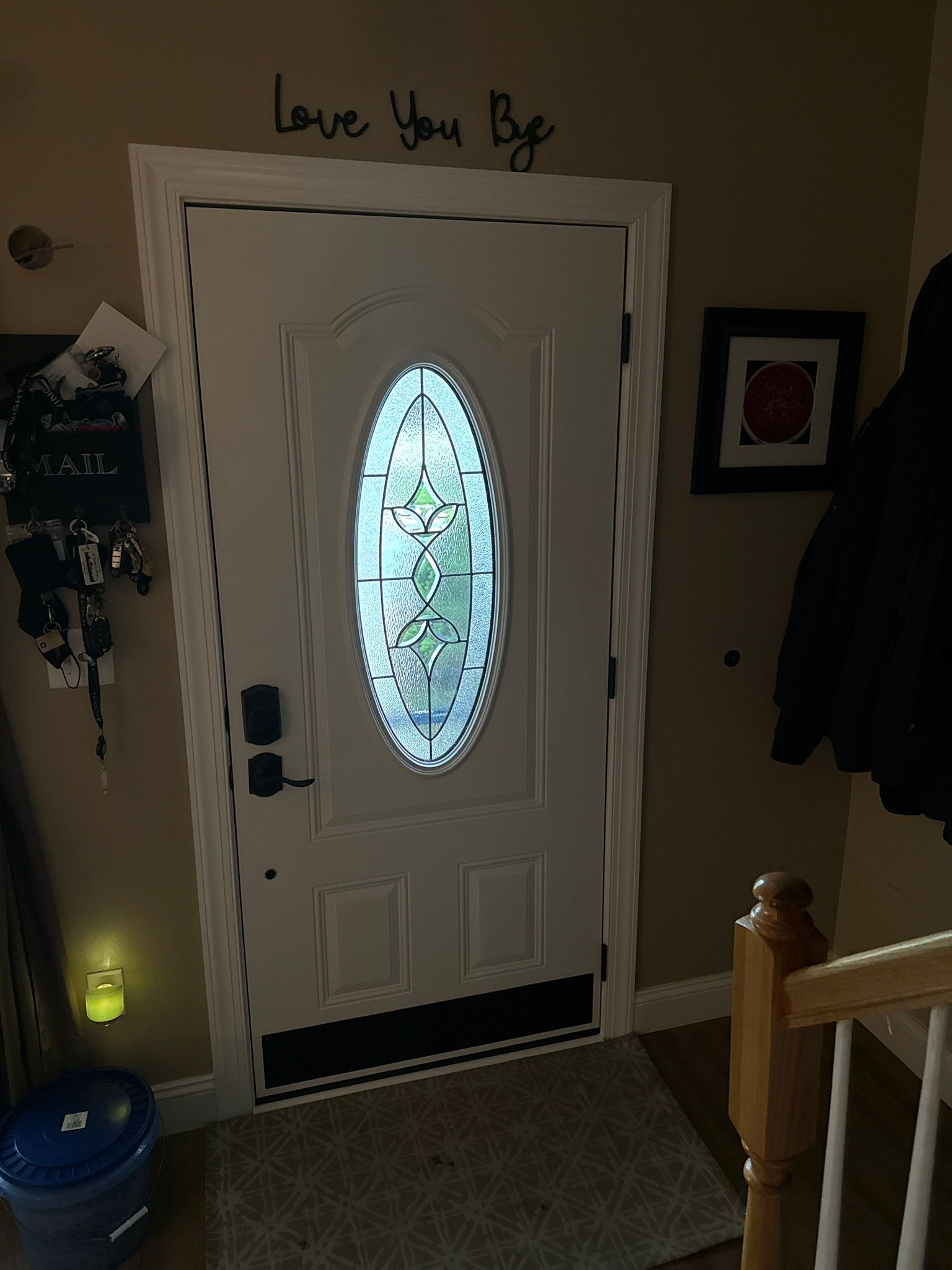Front door with oval frosted glass insert in a hallway, with wall decor and a stair railing nearby