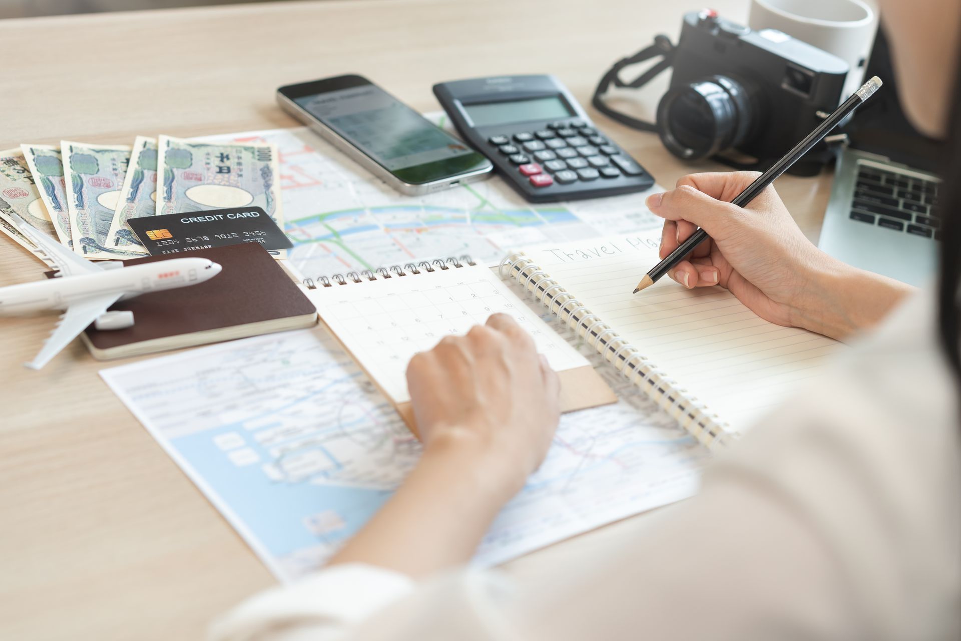 Person writing in a notebook at a desk with calculator, phone, camera, and papers