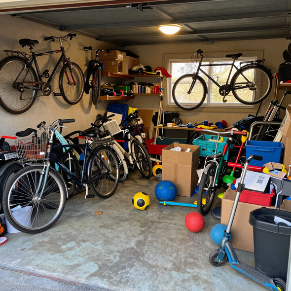 Cluttered garage packed with bicycles, scooters, boxes, and toys on a concrete floor.