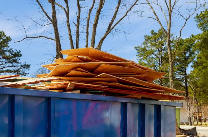 Pile of wooden planks in a blue dumpster, with trees and a blue sky in the background.