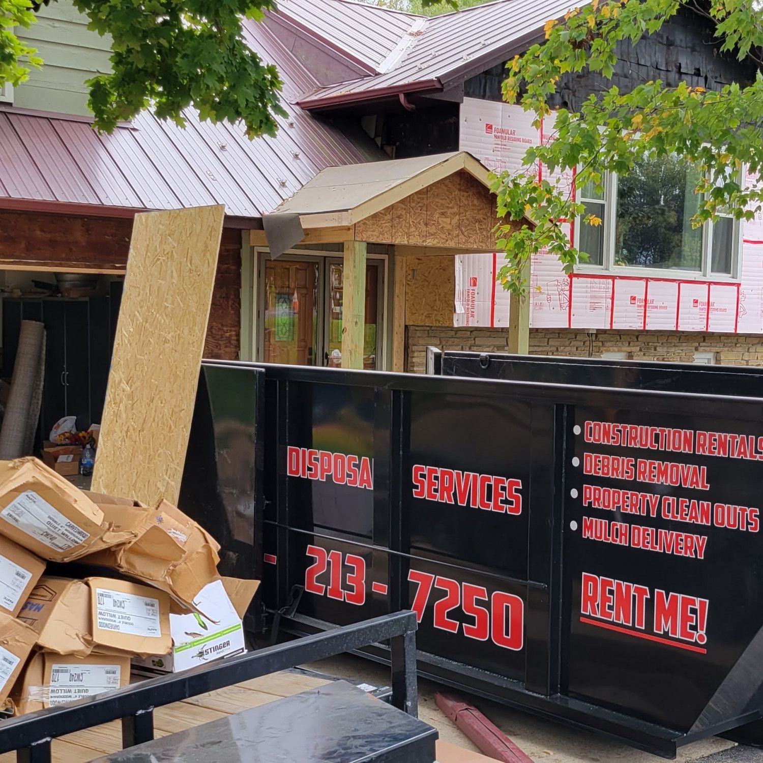 A black dumpster in front of a house under construction. Brown cardboard boxes are piled up nearby.