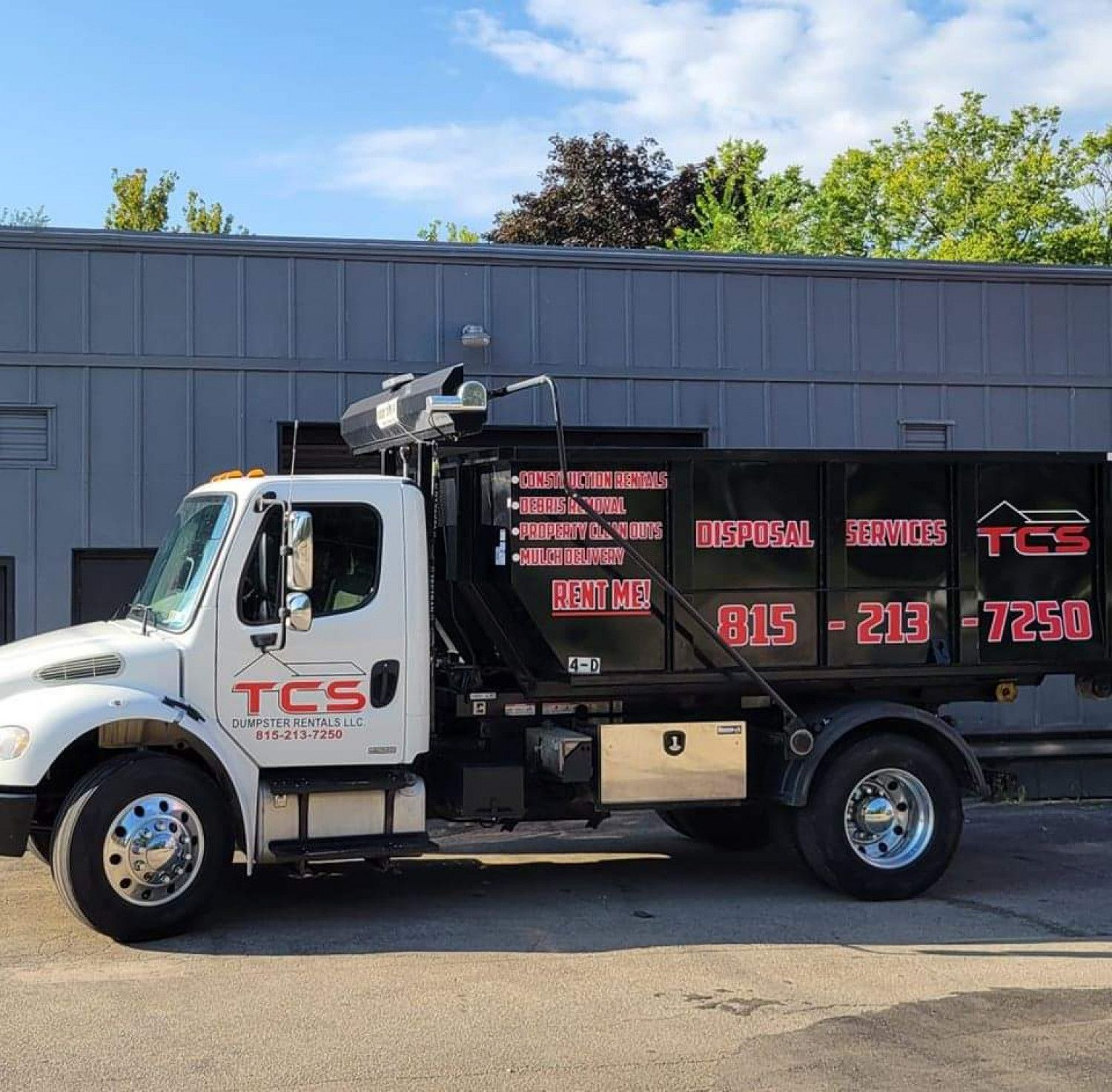 White TCS truck with a black dumpster bed parked in front of a gray building.
